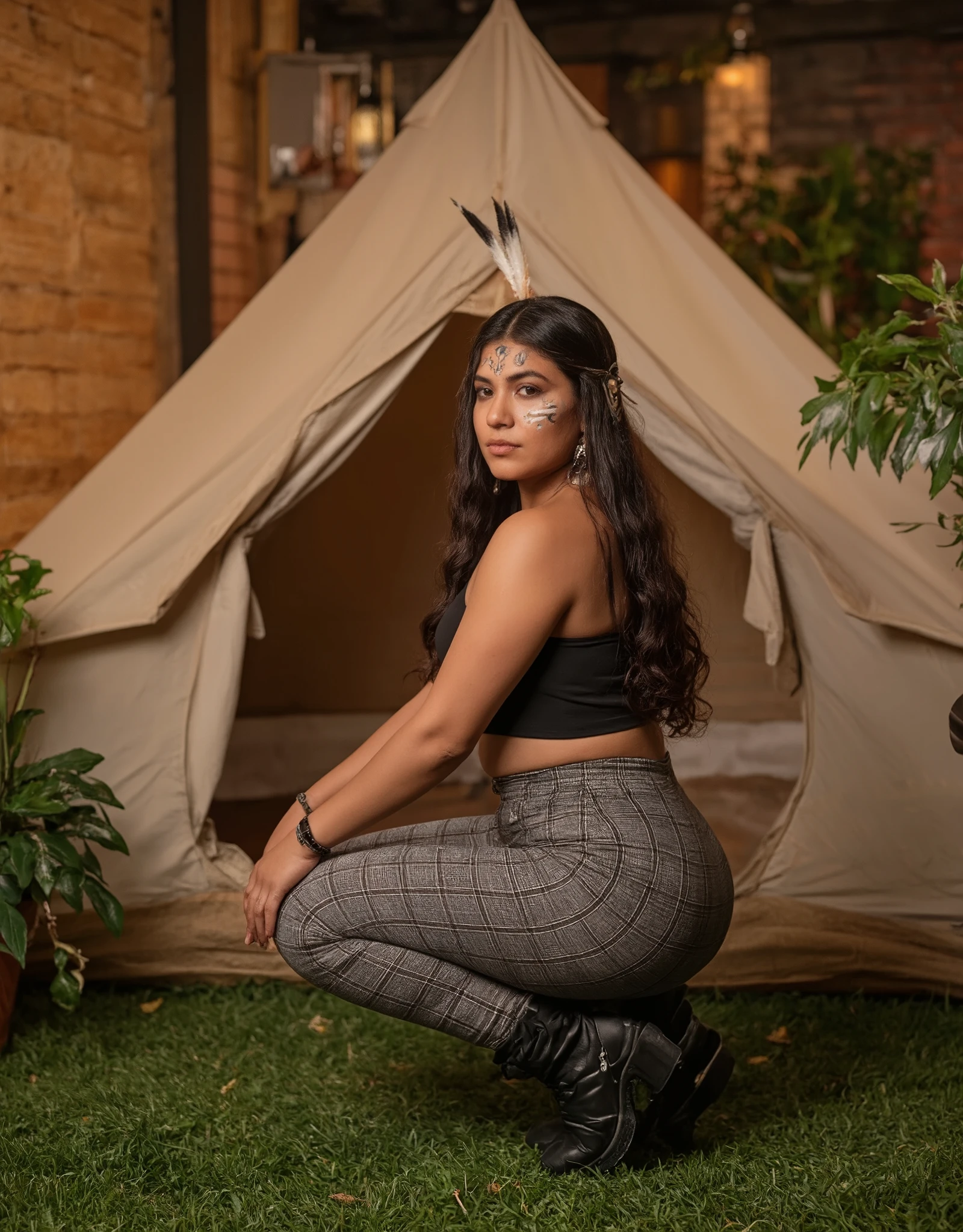 A professional photograph of a young woman with a curvy physique and long, wavy dark brown hair, posing in a stylish and confident stance. She is wearing a black fitted crop top and high-waisted gray plaid leggings that accentuate her figure. On her feet, she wears simple, comfortable open-toe sandals. She is squatting with her legs apart, her hands resting on her knees, and her back facing the camera, showcasing her posture. She is adorned with a vibrant indigenous headdress made of colorful feathers, adding a striking and cultural element to her look. Her face features traditional indigenous paint in bold, symbolic patterns, enhancing the authenticity and depth of her appearance. The background features a cozy indoor camping setup with a pitched tent on artificial grass, surrounded by wooden walls, exposed brick, and decorative plants. The lighting is warm and soft, casting a natural glow on her skin and creating a relaxed, inviting atmosphere. The textures of the wooden panels, brick wall, and fabric of the tent are clearly visible, adding depth and realism to the scene. The overall composition is well-balanced, with a high level of detail and a natural, immersive feel.
