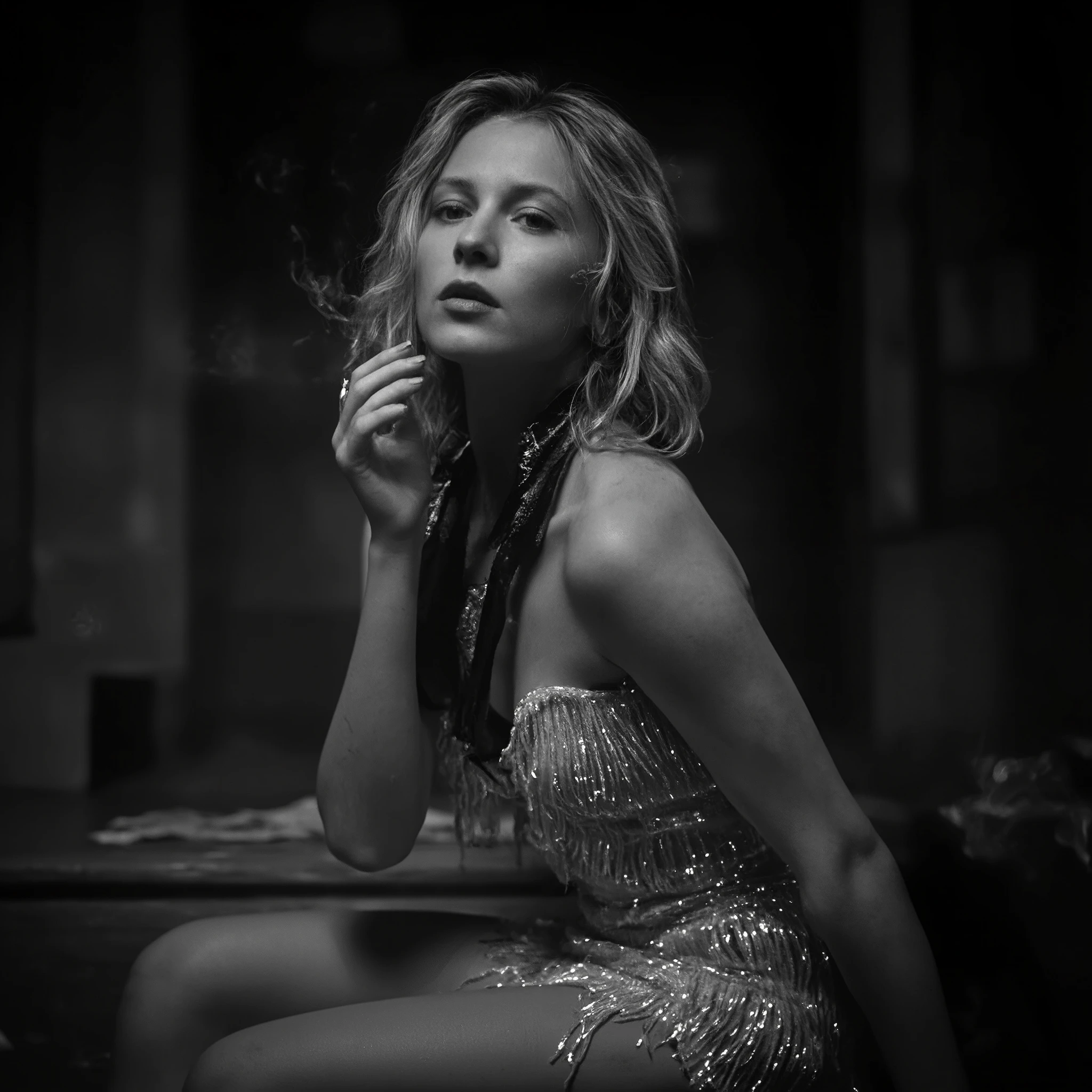 Create a cinematic close-up shot of a captivating blonde woman with loose strands of hair framing her face. She sits on a wooden staircase in a dimly lit space, wearing a shimmering fringed dress that catches the light. A dark scarf or necklace around her neck adds a touch of mystery. She holds a cigarette near her lips, with smoke curling in the air. Her gaze is distant and contemplative. Use high-contrast black and white tones to enhance the drama, with deep shadows and highlights sculpting her features. A blurred background creates depth, contributing to a raw and evocative atmosphere reminiscent of film noir aesthetics.
