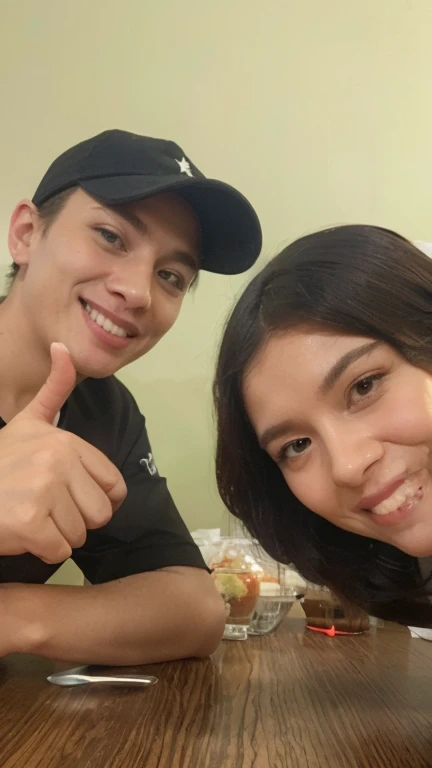 a couple taking a selfie in an indoor environment that appears to be a shopping mall food court. The woman is on the left, wearing a red blouse and smiling at the camera. The man is on the right, vestindo uma camiseta vermelha com uma assinatura preta, with a serious expression. Em primeiro plano, there is a yellow tray on the table, where there is also a cell phone with the screen on