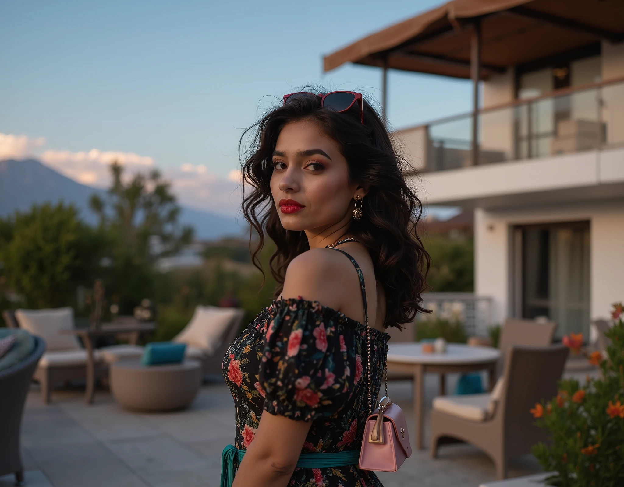 Photograph of a young, beautiful Indian woman with shoulder-length dark brown wavy  hair and pale white skin, wearing bright lipstick. She has a heart-shaped face and big brown eyes, dressed in a short black dress with colorful floral patterns and a teal waistband. She accessorizes with a small, light pink handbag and sunglasses perched on her head. She stands against the serene backdrop of a modern outdoor patio area at dusk. The circular wicker table and matching chairs are visible in the foreground, with a small tree to the right. In the background, a two-story white building with a large brown canopy covers the upper deck, and the sky transitions from blue to a warm, soft hue near the horizon, adding to the tranquil atmosphere.