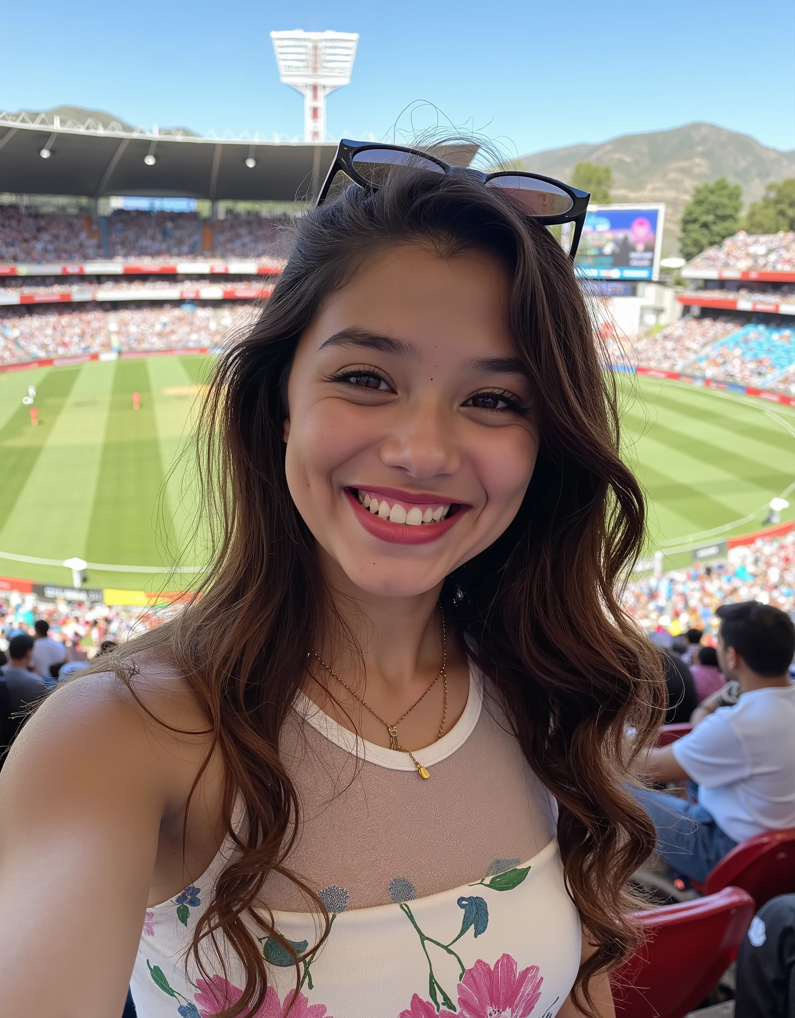 A selfie photo of a young beautiful indian woman with long, wavy brown hair and a fair complexion, pale white skin, dimple on cheeks, smiling widely at the camera. She has dark eyebrows, brown eyes, and is wearing pink lipstick. She is wearing sunglasses on her head and a gold necklace.white floral sheer crop top. The background shows a packed stadium with red seats and a green field in the center, where a cricket match is in progress. The crowd is densely packed, and the stadium is brightly lit, indicating a daytime match. The image is vibrant and dynamic, capturing the energy of the event.