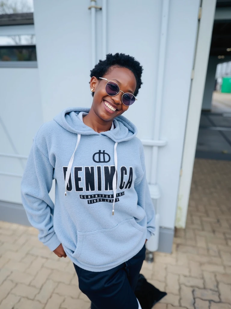 (photorealism:1.2), beautiful black African woman,22 years,sharp boobs,,wearing jeans and a fitting white hoodie written "Kinuthia", smiling,long braids,oversized blue photo chromatic glasses, standing , plants in background, Nickie Minaj inspiration