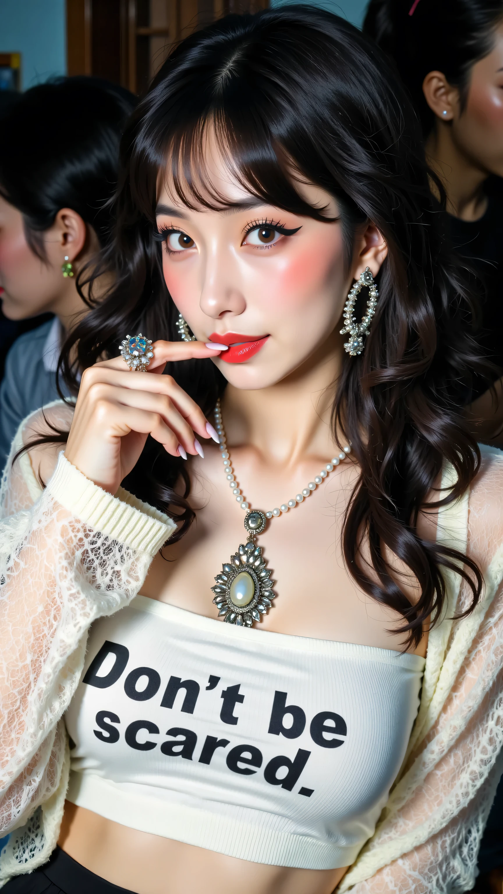 8k, Masterpiece, Top Quality, Close-up, slightly high angle viewpoint, woman with dark, voluminous wavy hair with bangs, fair skin, light eyes with dark eyeliner, red lipstick, wearing a white cropped top with black text ("Don't be scared"), a sheer white long-sleeved cardigan, a pearl necklace with a large ornate pendant, and large silver hoop earrings, one hand near her mouth with long nails, winking, looking towards the camera with a playful expression, blurred background suggesting an indoor setting with dark tones.