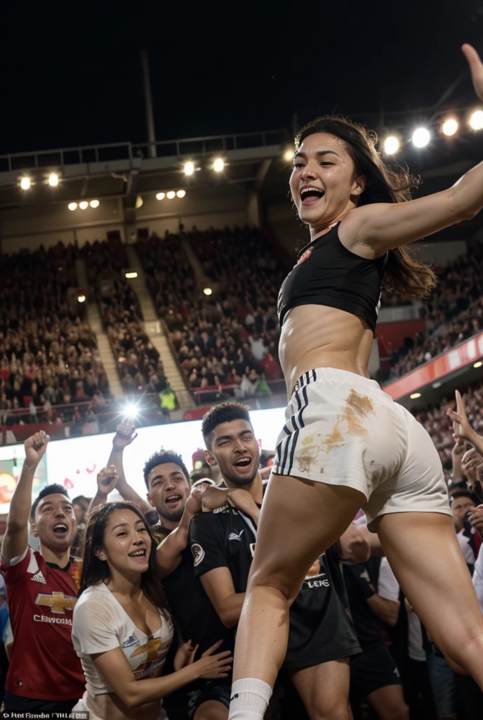 Athletic attractive asian woman with muscular arms wearing sports bra and shorts kneeling on ground celebrating winning beach wrestling match, fists in air. Her shirtless male opponent lying next to her injured and crying in pain, blood on face and chest. Crowd in background