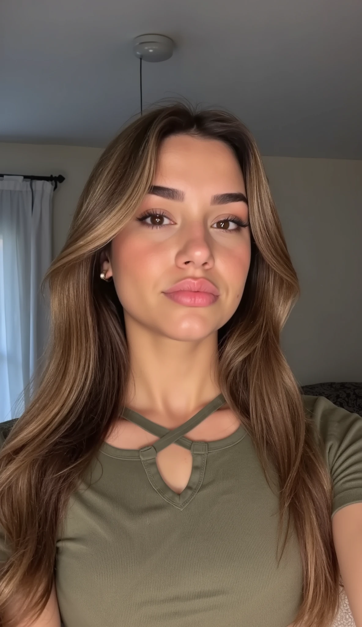 Realistic daytime portrait photo of Mikayla, a young attractive woman with a calm and neutral expression, standing in a softly lit room. She has long, straight caramel-blonde hair with a natural center part. Her skin is smooth and glowing with subtle natural makeup, including soft brows, a light touch of mascara, and nude glossy lips. She is wearing a casual olive green top with a subtle cut-out detail at the neckline. The background is minimalistic, featuring a soft gray wall and parts of a white curtain, softly blurred to keep the focus on her. Natural light from a window illuminates her face evenly, creating a bright and authentic home atmosphere. iPhone-style composition, realistic and clean aesthetic, perfect for a casual story post.