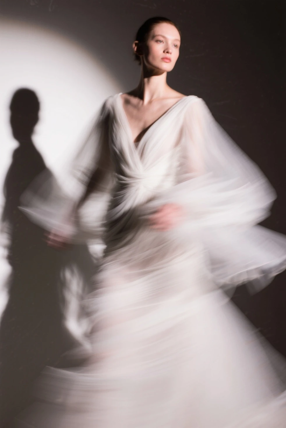 Realistic photographic image: On a top model catwalk, enveloped in dry ice vapors. view from side, with delicate and decisive movements. A tall, slender woman; approximately 28 years old; caucasiana; platinum hair styled in a low bun on the nape of her neck; wearing flexible clothing, loose to shapes and movements; wearing matching high heels. she is elegant, modern and futuristic with robotic details in the form of delicate golden jewelry; with innovative aesthetic posture, evaluated and approved by the academic criteria adopted by critics, beauticians, advertising and visual arts curators. The high contrast between the deep black background and the texture of the art evokes a feeling of evolutionary fullness.