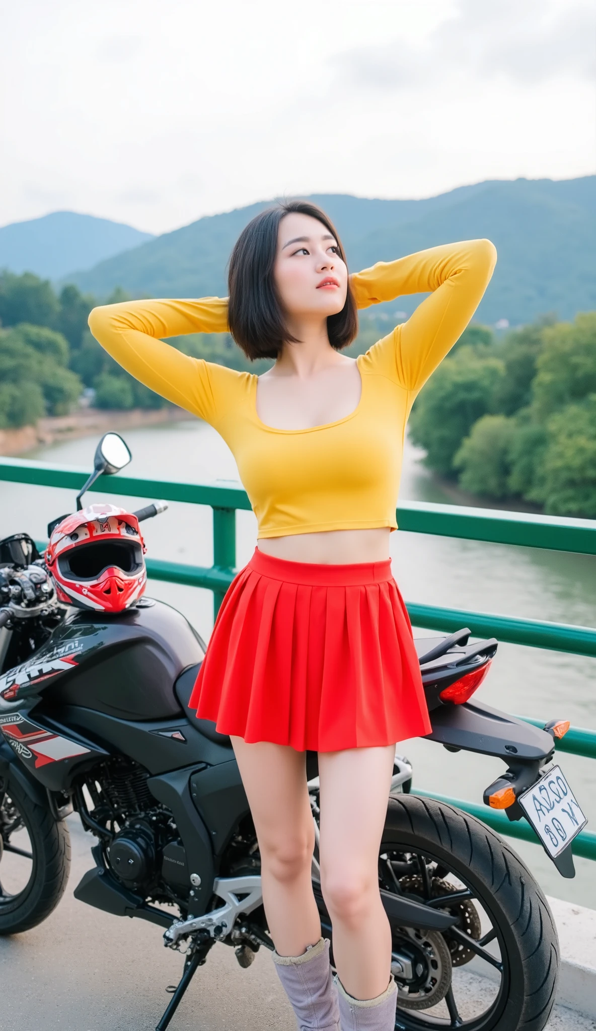 A young Thai woman has a good figure curve and large breasts with short dark hair, wearing a yellow long-sleeved top, a short red pleated mini skirt, light-colored stockings, and colours racing boots. She is standing next to a Suzuki motorcycle style touring on a bridge or elevated walkway, looking up and to the side with her arms raised behind her head. A motorcross helmet style rests on the motorcycle. The background shows a landscape with a river, trees, and hills under a slightly overcast sky.