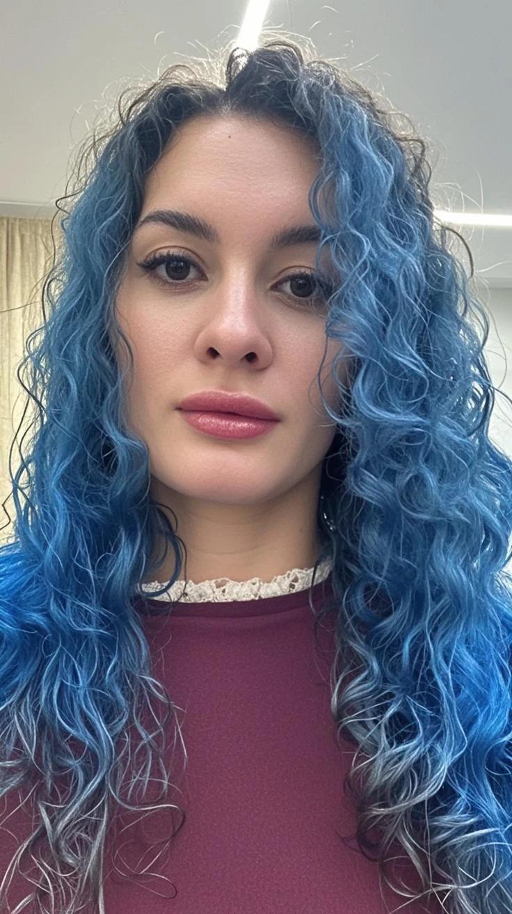 A young woman with blue hair styled in loose waves, looking off to her right with a subtle smile. She is wearing a light bluish blazer over a black top and has a delicate necklace around her neck. The background suggests an indoor setting with neutral tones