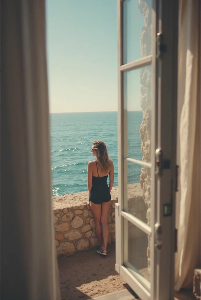 A photograph capturing a serene moment of a nude woman standing leaning against an open double door, looking out towards the ocean. The woman is seen from behind, with her right arm raised and touching the right door jamb, while her left arm hangs naturally at her side. Her long, wavy hair flows down her back, creating a silhouette against the bright background. The doors, white with glass panels on either side, are slightly ajar, offering a clear view through the central opening. The interior room is dimly lit, creating a stark contrast to the bright exterior. To the left of the frame, there is a partial view of a wooden chair with a curved back and slender legs, while to the right, part of a sofa is visible. The interior floor is made of dark tiles arranged in a herringbone pattern, reflecting some light. Through the balcony railing, which features elaborate black wrought-iron scrollwork and white balustrades, a terracotta tiled surface is visible. On the left side of the balcony, a wooden chair with a curved back and thin legs faces the ocean. The background shows a tranquil seascape with calm waters stretching to the horizon, meeting a sky that changes from a pale peach near the horizon to azure blue higher up.