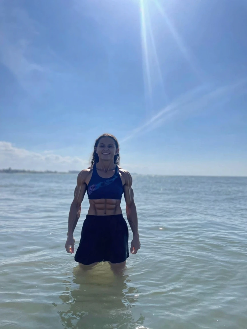 Front angle portrait, highly detailed RAW color photo, 4k, front angle, full body, toned body, muscular, wearing black metallic suplex swimsuit, floss suit, standing in water, large breasts, fitness body, is wearing swimsuit type metallic armor, big chest, floss style, floss model, beautiful girl, profile picture, profile picture, full body, large breasts, round breasts, on the beach, in the water up to the feet, Brazilian, (looking up), toned body, highly detailed, sunset lighting,