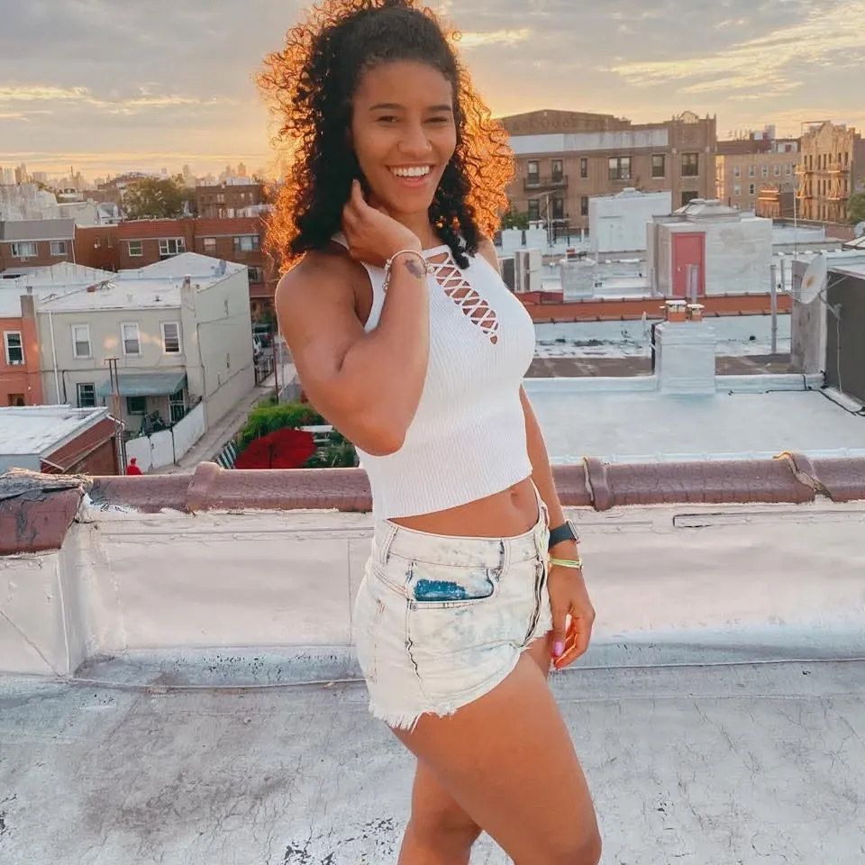 a beautiful biracial woman, blue hair, light blue eyes, athletic figure, large breasts, extremely detailed full body, sunset, on a balcony, drink in the hand, light of sun reflect on face, yellow t-shirt, jeans skirt