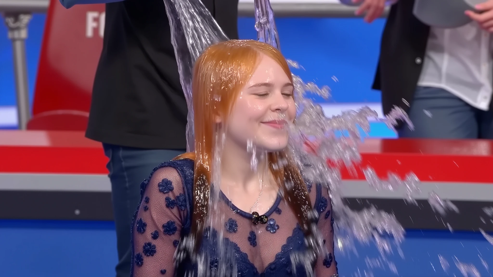 On a gameshow, a beautiful young 18-year-old girl with tightly curly ginger hair and wearing a dark blue lace top dress with puff sleeves, a thin silver necklace, makeup including lipstick and eye shadow, long false eyelashes, and long press-on false nails has a bucket of water poured into her face from off screen. The water lands in the middle of her face and spreads out, completely soaking her eyes, nose and mouth. Photorealistic. 1girl. Solo. Medium close-up. Angled view.
