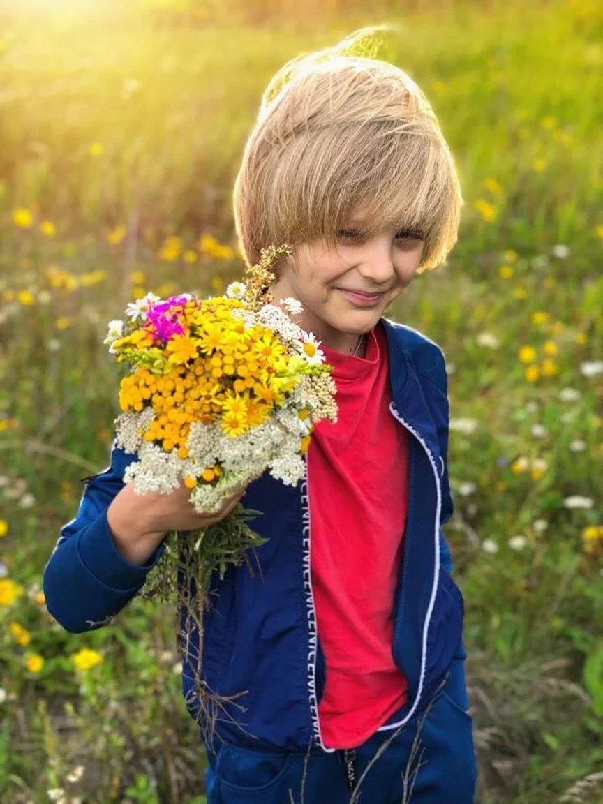 A Russian boy with blond hair in a white long-sleeved shirt and olive pants stands near a blooming apple tree and looks up