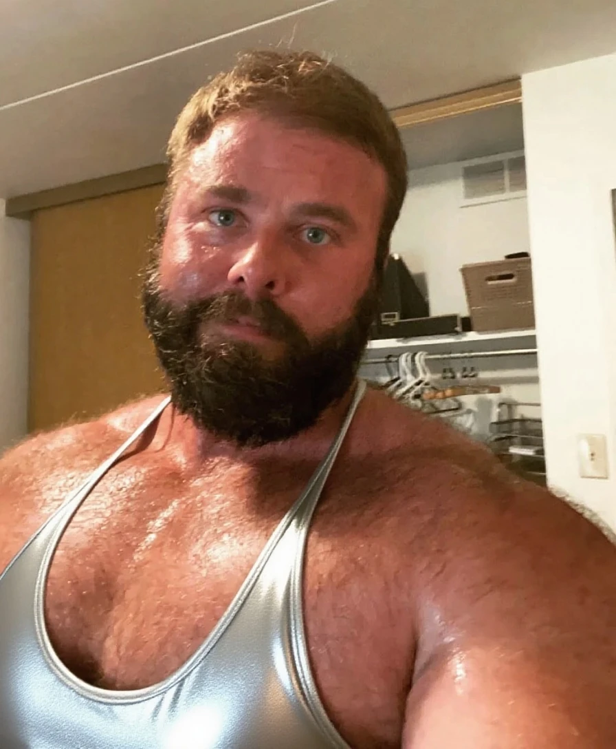 Foto de um homem de 40 anos, German and white with light, pink skin, vestindo uma camiseta regata branca e usando um short curto de cor vermelha. Tem barba curta e cabelo curto, his chest is robust and his body is muscular, he also has a little belly. He is in a relaxed and carefree pose, the lighting is soft and natural, lembrando fotografias profissionais. The man looks like a daddy and is cute and sensual at the same time
