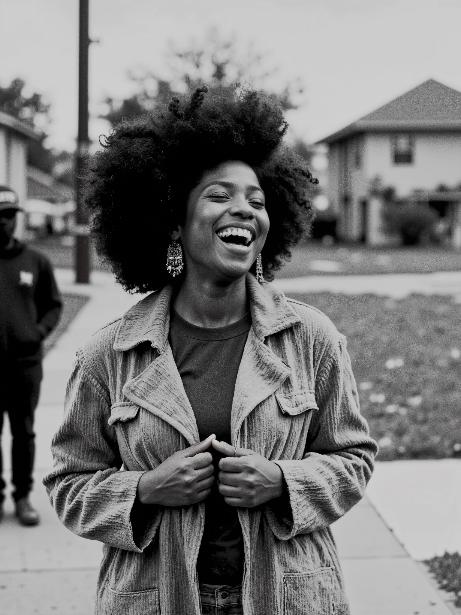 Ultra-realistic black and white photograph of a joyful woman laughing heartily in an outdoor urban neighborhood setting. She has a voluminous curly afro hairstyle and wears large, ornate chandelier earrings. Her expression is full of life and energy, mouth wide open in mid-laugh, eyes nearly closed from the intensity of her joy. She is dressed in a textured, vintage-style corduroy trench coat layered over a dark shirt, gripping the coat near her chest with one hand. The background features blurred figures and residential houses, adding depth and context to the scene. The image captures raw emotion with rich texture details in the clothing, hair, and skin, embodying a candid and vibrant moment of happiness.