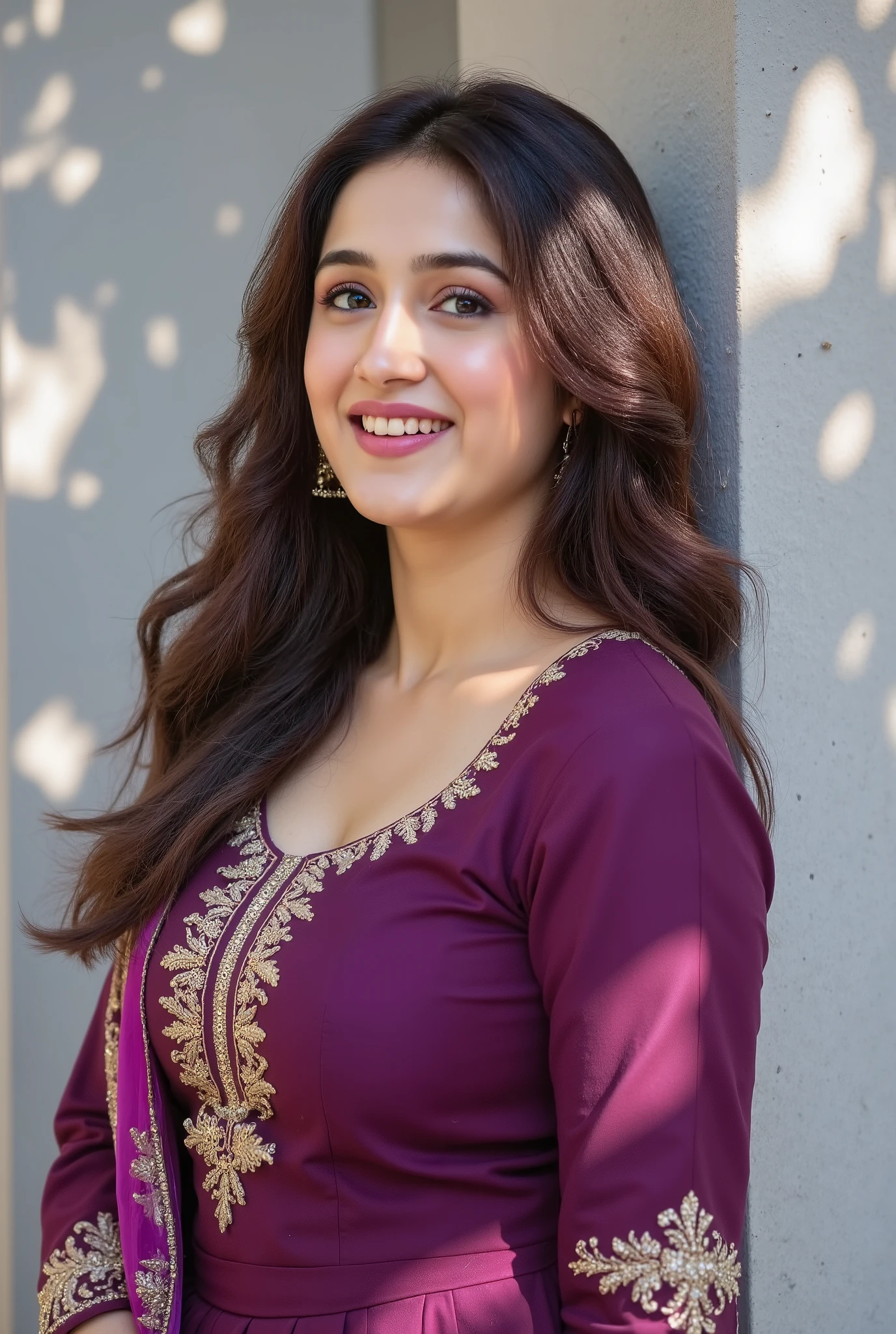 A young woman with long wavy hair smiles warmly while leaning against a textured light grey wall under dappled sunlight and shadow from leaves. She is wearing a deep purple traditional Indian outfit with intricate gold embroidery on the front, and a matching dupatta draped elegantly. Her natural makeup, small bindi, and joyful expression create a serene, candid, and elegant mood. The composition is bright, airy, and minimal, capturing the warmth of a sunlit moment with soft, natural shadows and highlight