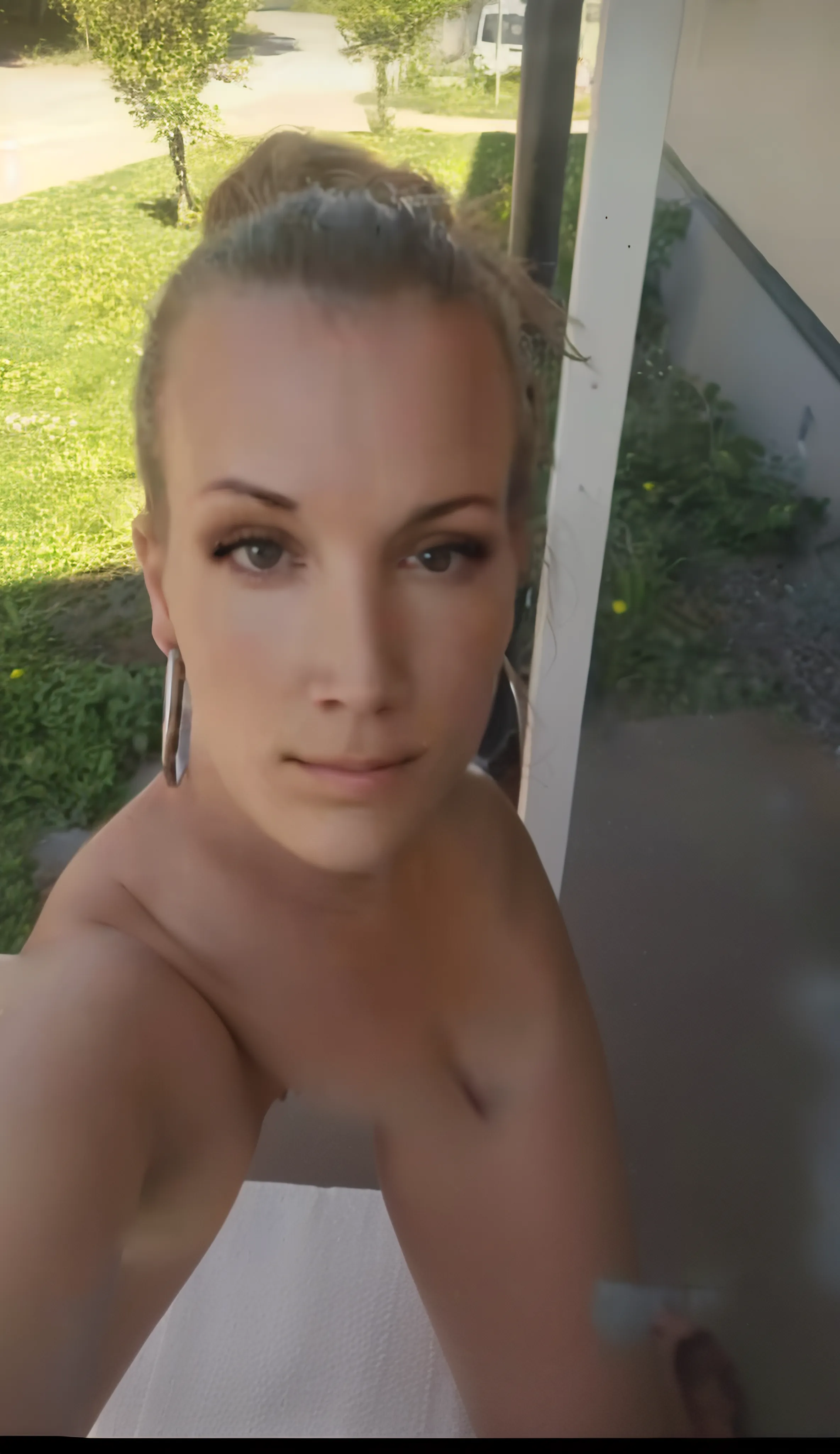A blonde woman in a very sexy selfie of her face on a balcony where behind we see a beach 