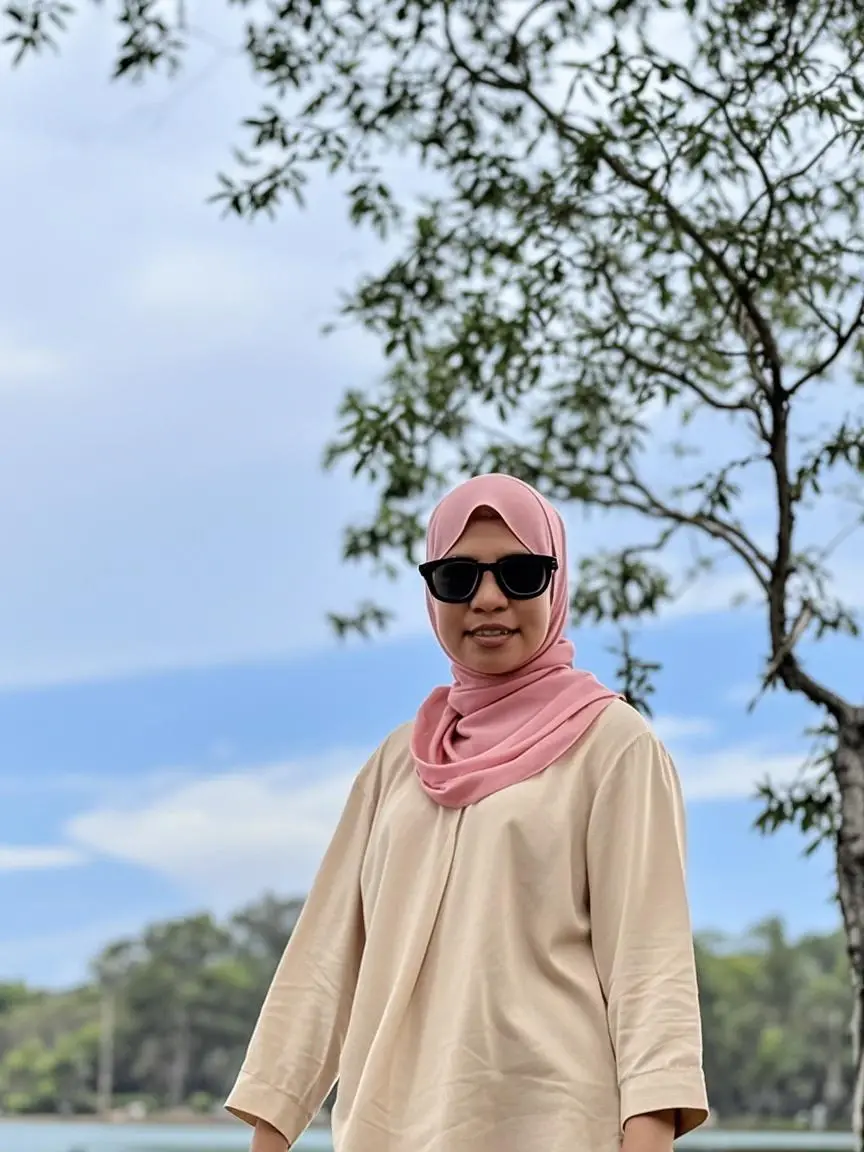 A beautiful young Muslim woman wearing a long hijab and glasses with a loose white t-shirt that says I Love Palembang, sandy beach and rock cliffs in the background, wide angle view full shot