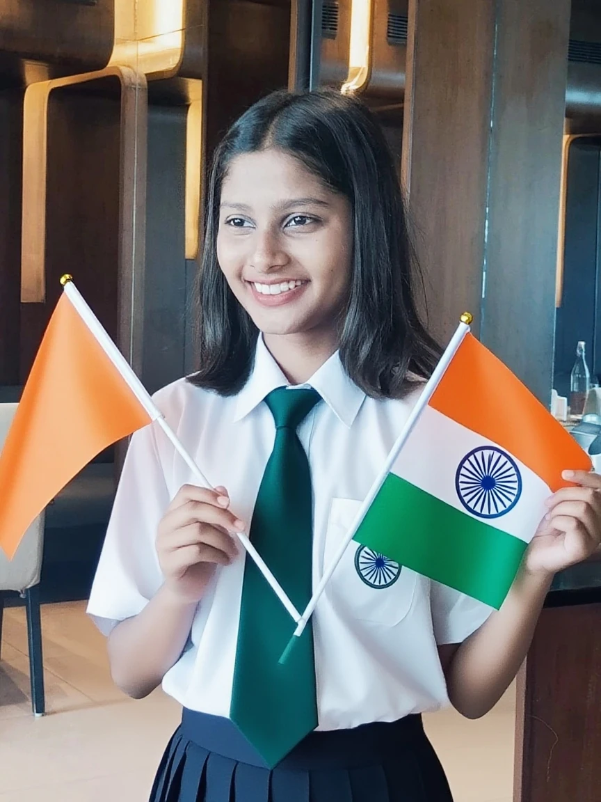 4 year old girl is holding Indian flag in her hand, she is wearing white kurti or black leggings, there is school or school board in the background, name of Jishpar school is written as silver oak international school