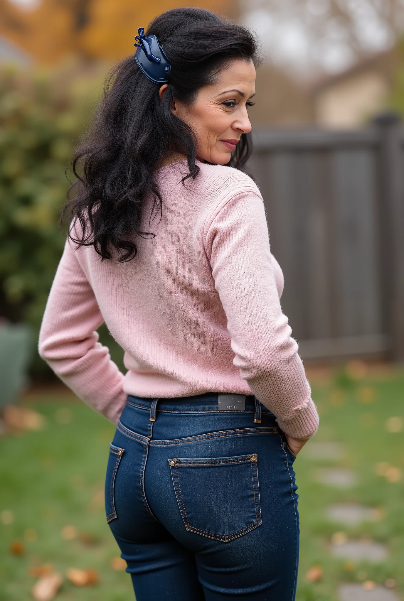 hyperrealistic full-body true rear portrait of mature European woman with unmistakable mom presence captured directly from behind with perfect vertical alignment, long dark wavy hair with natural texture variation gathered and secured with dark blue plastic claw clip allowing elegant tendrils to escape framing neck area, head turned precisely at 45-degree angle over right shoulder revealing partial profile with graceful mature beauty showing refined features and subtle signs of aging adding character, wearing ultra-skinny dark blue jeans with exceptional second-skin fit containing high elastane percentage creating perfect fabric tension across wide pear-shaped buttocks with characteristic pocket placement highlighting wide hip structure contrasting dramatically with notably slim waist, pronounced hourglass body shape with significant proportional difference between hip and waist measurements creating classical feminine silhouette, pale pastel rose form-fitting knit sweater with authentic textile weave showing subtle ribbing pattern at cuffs and hemline, deliberately tucked slightly into waistband at back creating emphasis on waistline and enhancing natural curves, standing with weight slightly shifted to one leg creating subtle natural asymmetry in quiet secluded backyard area with minimal landscaping elements, soft blurred background with muted autumn foliage creating professional bokeh effect enhancing subject isolation, captured in diffused overcast autumn daylight creating even illumination with subtle directional quality, cinematic ultra-detailed 8k photography style with perfect technical execution