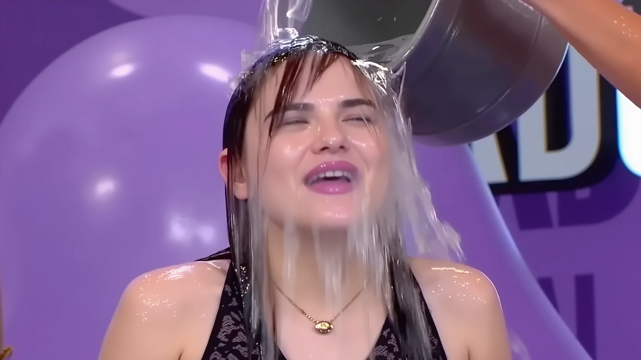 On a gameshow, a beautiful young 18-year-old girl with long purple hair and wearing a black lace dress through which her black lace bra can be seen, a thin gold necklace, makeup including purple lipstick and purple eye shadow, long false eyelashes, and long purple press-on false nails has a bucket of water poured into her face from off screen. The water lands in the middle of her face and spreads out, completely soaking her eyes, nose and mouth. Photorealistic. 1girl. Solo. Close-up.