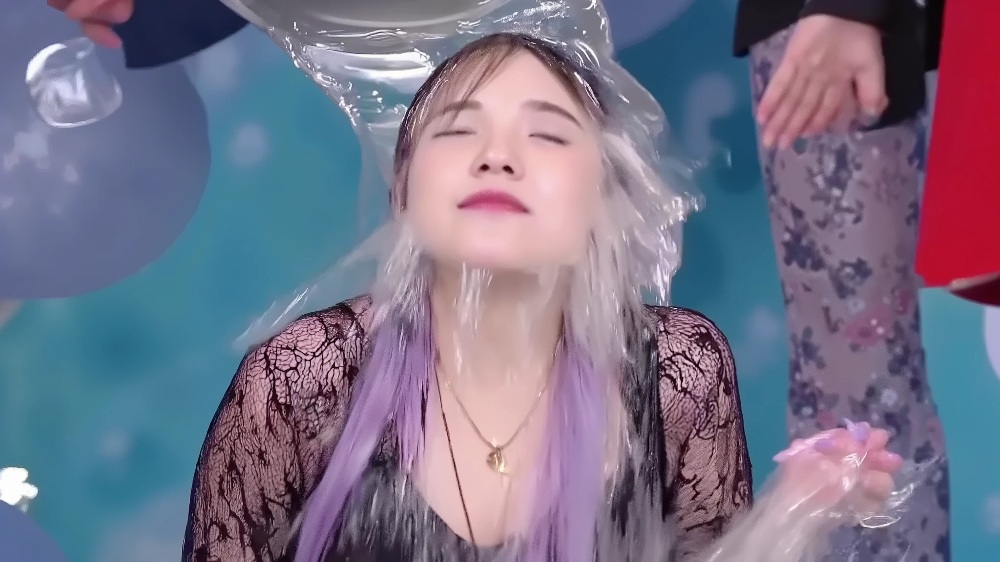 On a gameshow, a beautiful young 18-year-old girl with long purple hair and wearing a black lace dress through which her black lace bra can be seen, a thin gold necklace, makeup including purple lipstick and purple eye shadow, long false eyelashes, and long purple press-on false nails has a bucket of water poured into her face from off screen. The water lands in the middle of her face and spreads out, completely soaking her eyes, nose and mouth. Photorealistic. 1girl. Solo. Close-up.