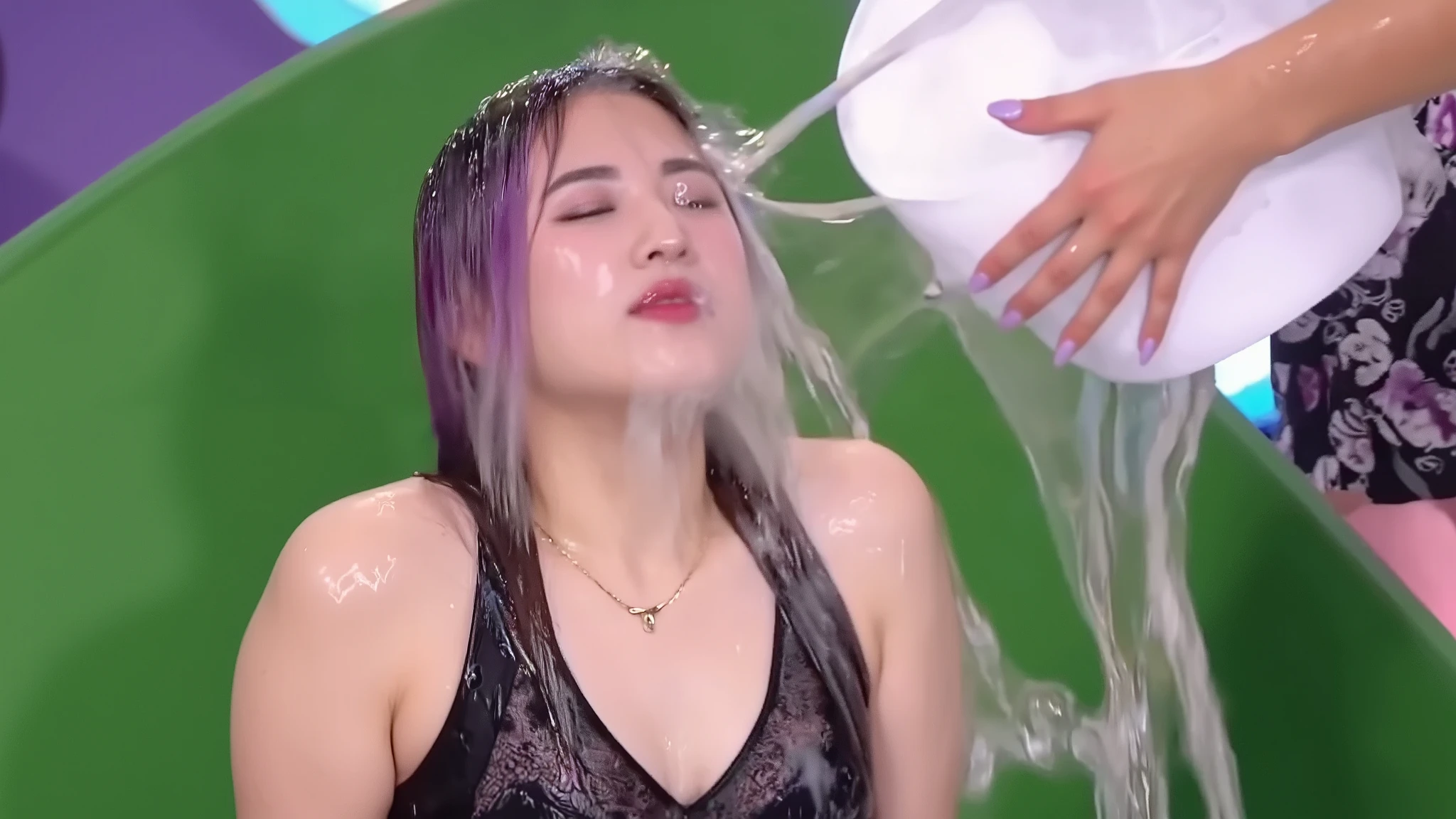 On a gameshow, a beautiful young 18-year-old girl with long purple hair and wearing a black lace dress through which her black lace bra can be seen, a thin gold necklace, makeup including purple lipstick and purple eye shadow, long false eyelashes, and long purple press-on false nails has a bucket of water poured into her face from off screen. The water lands in the middle of her face and spreads out, completely soaking her eyes, nose and mouth. Photorealistic. 1girl. Solo. Close-up.
