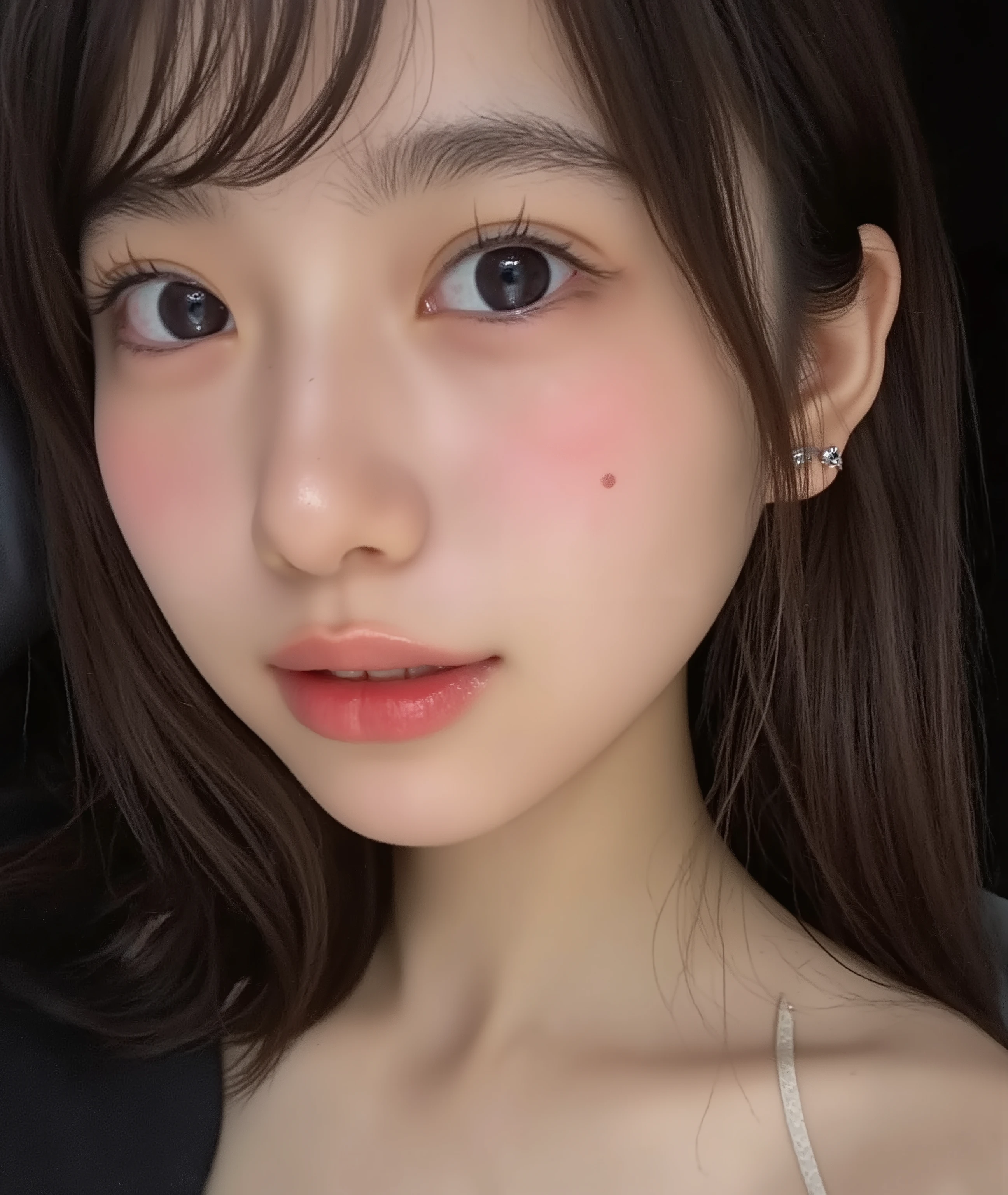The image is a close-up photograph of a young Asian woman with a fair complexion. Her face dominates the frame, occupying the majority of the image, with her eyes, nose, and lips prominently visible. She has large, almond-shaped eyes with long, thick eyelashes, and her skin is smooth and soft. Her lips are full and slightly parted, revealing a natural, rosy hue. She wears a small silver stud earring in her left ear, adding a subtle touch of personal style.

Her hair is long, dark brown with subtle highlights, and it partially covers her forehead, giving a slightly tousled, casual look. The background is out of focus, but it appears to be a dark, possibly indoor setting, which emphasizes her face and provides a stark contrast to her light complexion. She is wearing a black garment, possibly a top or a sleeve, which is visible at the bottom of the image. The overall mood of the photograph is intimate and personal, capturing a candid moment with a natural, unfiltered aesthetic.