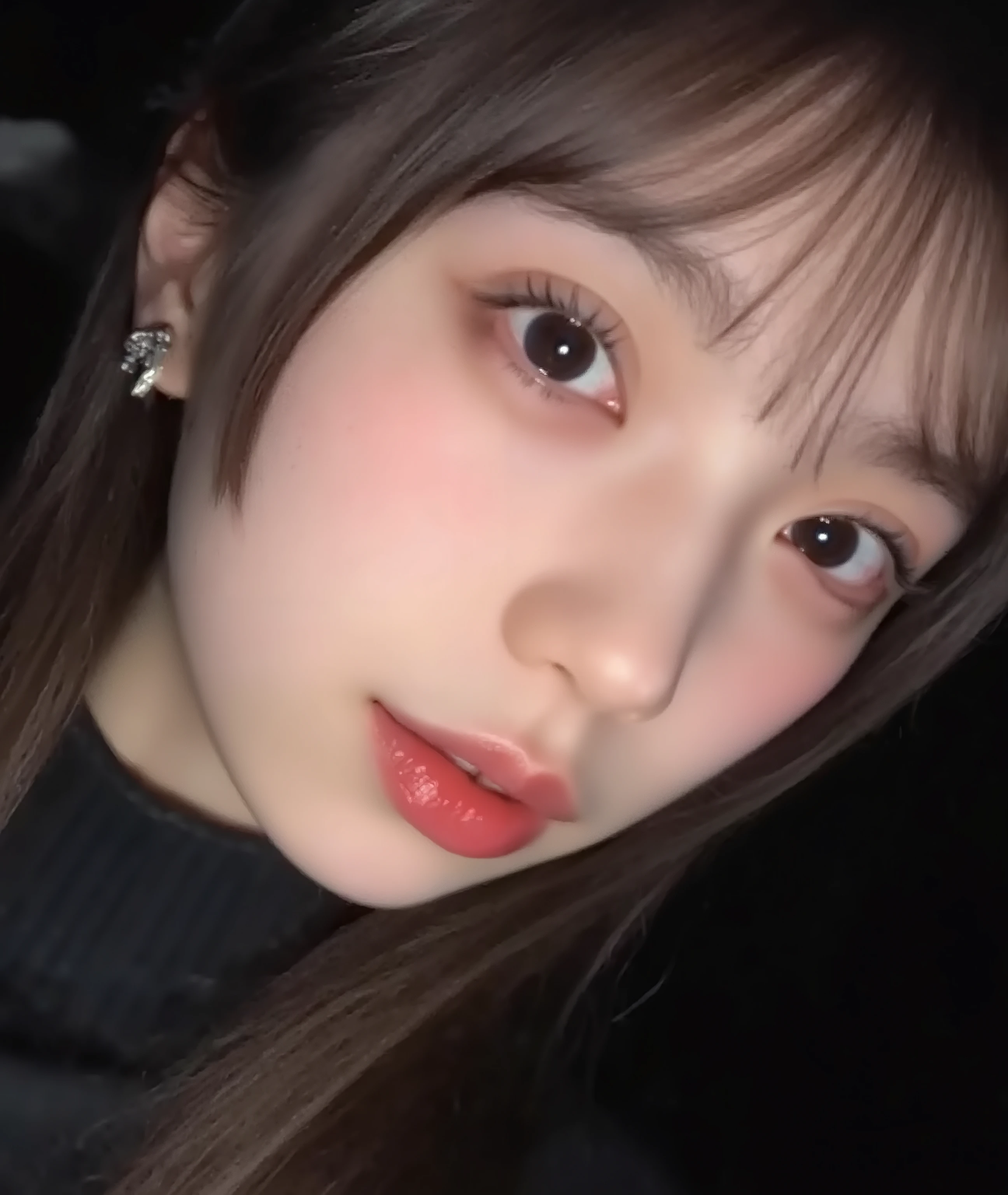 The image is a close-up photograph of a young Asian woman with a fair complexion. Her face dominates the frame, occupying the majority of the image, with her eyes, nose, and lips prominently visible. She has large, almond-shaped eyes with long, thick eyelashes, and her skin is smooth and soft. Her lips are full and slightly parted, revealing a natural, rosy hue. She wears a small silver stud earring in her left ear, adding a subtle touch of personal style.
Her hair is long, dark brown with subtle highlights, and it partially covers her forehead, giving a slightly tousled, casual look. The background is out of focus, but it appears to be a dark, possibly indoor setting, which emphasizes her face and provides a stark contrast to her light complexion. She is wearing a black garment, possibly a top or a sleeve, which is visible at the bottom of the image. The overall mood of the photograph is intimate and personal, capturing a candid moment with a natural, unfiltered aesthetic.