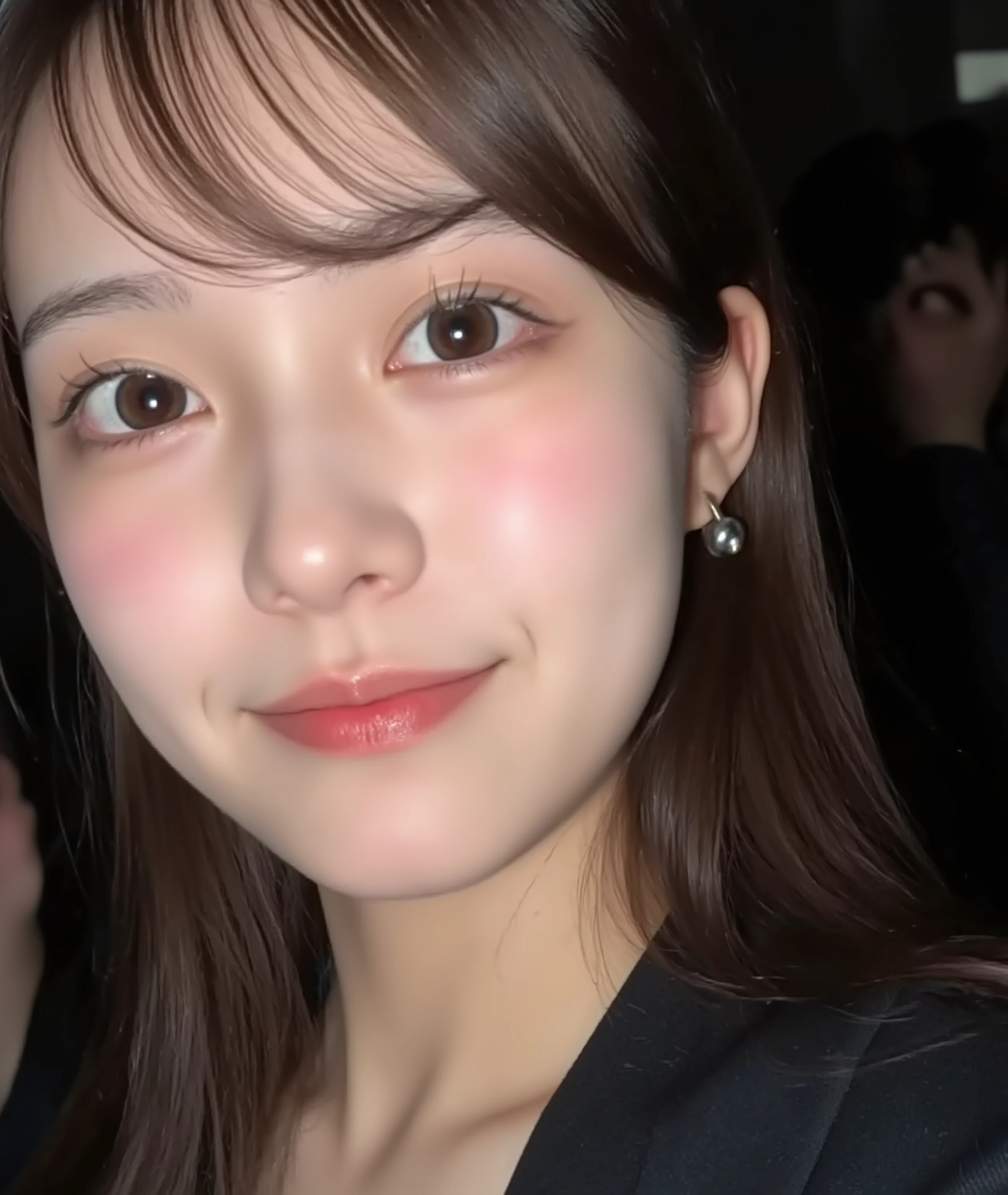 The image is a close-up photograph of a young Asian woman with a fair complexion. Her face dominates the frame, occupying the majority of the image, with her eyes, nose, and lips prominently visible. She has large, almond-shaped eyes with long, thick eyelashes, and her skin is smooth and soft. Her lips are full and slightly parted, revealing a natural, rosy hue. She wears a small silver stud earring in her left ear, adding a subtle touch of personal style.
Her hair is long, dark brown with subtle highlights, and it partially covers her forehead, giving a slightly tousled, casual look. The background is out of focus, but it appears to be a dark, possibly indoor setting, which emphasizes her face and provides a stark contrast to her light complexion. She is wearing a black garment, possibly a top or a sleeve, which is visible at the bottom of the image. The overall mood of the photograph is intimate and personal, capturing a candid moment with a natural, unfiltered aesthetic.