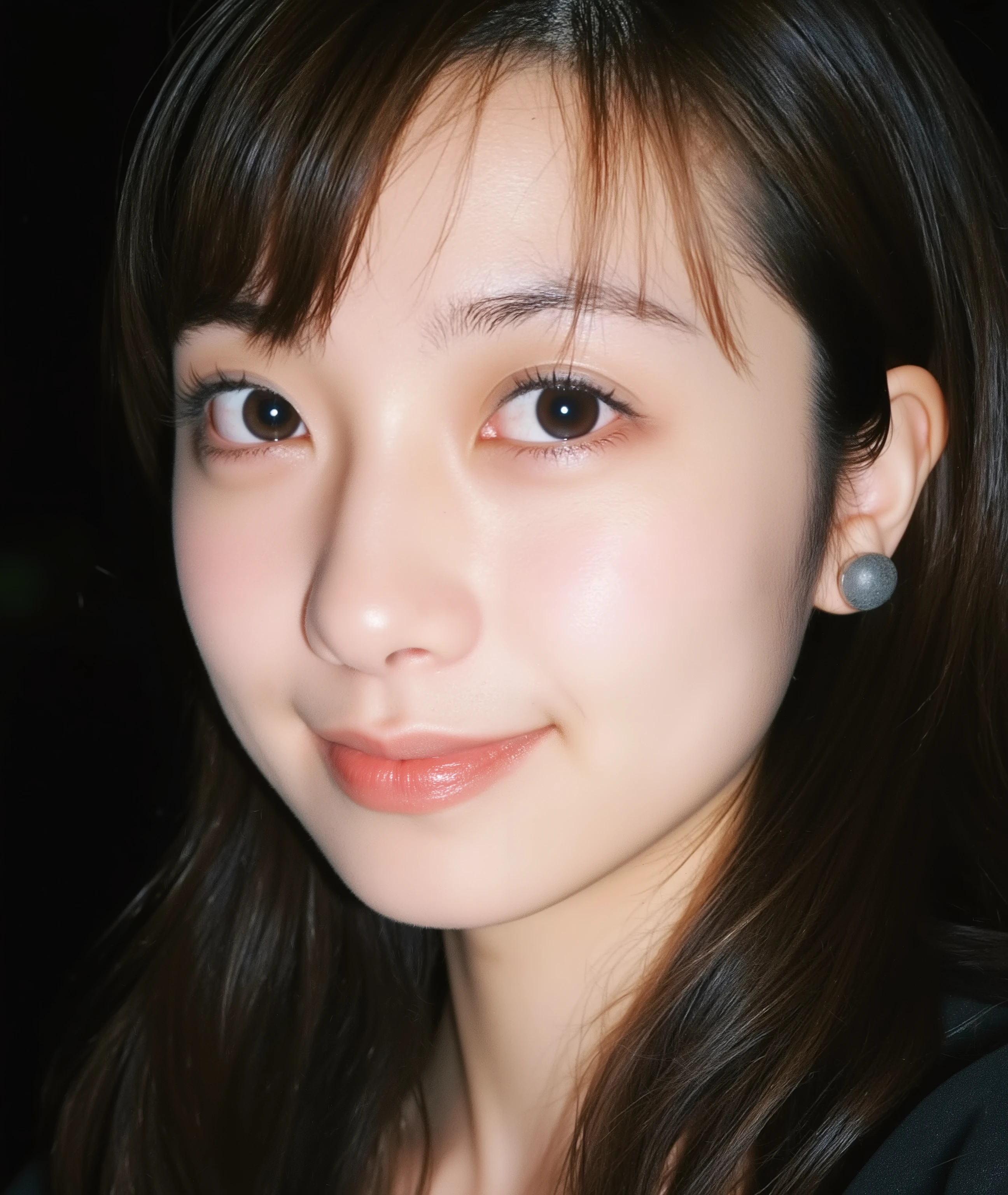 The image is a close-up photograph of a young Asian woman with a fair complexion. Her face dominates the frame, occupying the majority of the image, with her eyes, nose, and lips prominently visible. She has large, almond-shaped eyes with long, thick eyelashes, and her skin is smooth and soft. Her lips are full and slightly parted, revealing a natural, rosy hue. She wears a small silver stud earring in her left ear, adding a subtle touch of personal style.

Her hair is long, dark brown with subtle highlights, and it partially covers her forehead, giving a slightly tousled, casual look. The background is out of focus, but it appears to be a dark, possibly indoor setting, which emphasizes her face and provides a stark contrast to her light complexion. She is wearing a black garment, possibly a top or a sleeve, which is visible at the bottom of the image. The overall mood of the photograph is intimate and personal, capturing a candid moment with a natural, unfiltered aesthetic.