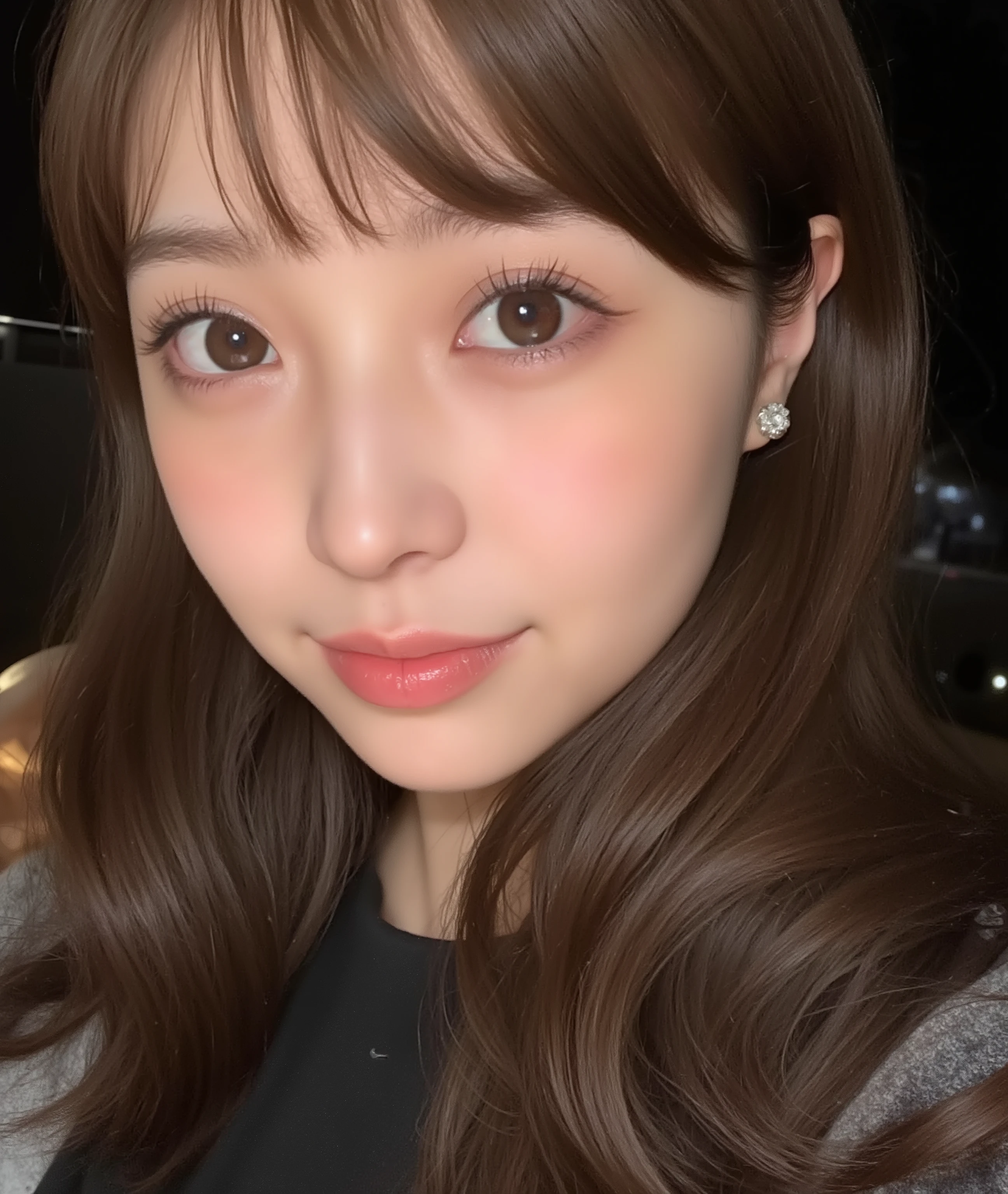 The image is a close-up photograph of a young Asian woman with a fair complexion. Her face dominates the frame, occupying the majority of the image, with her eyes, nose, and lips prominently visible. She has large, almond-shaped eyes with long, thick eyelashes, and her skin is smooth and soft. Her lips are full and slightly parted, revealing a natural, rosy hue. She wears a small silver stud earring in her left ear, adding a subtle touch of personal style.

Her hair is long, dark brown with subtle highlights, and it partially covers her forehead, giving a slightly tousled, casual look. The background is out of focus, but it appears to be a dark, possibly indoor setting, which emphasizes her face and provides a stark contrast to her light complexion. She is wearing a black garment, possibly a top or a sleeve, which is visible at the bottom of the image. The overall mood of the photograph is intimate and personal, capturing a candid moment with a natural, unfiltered aesthetic.