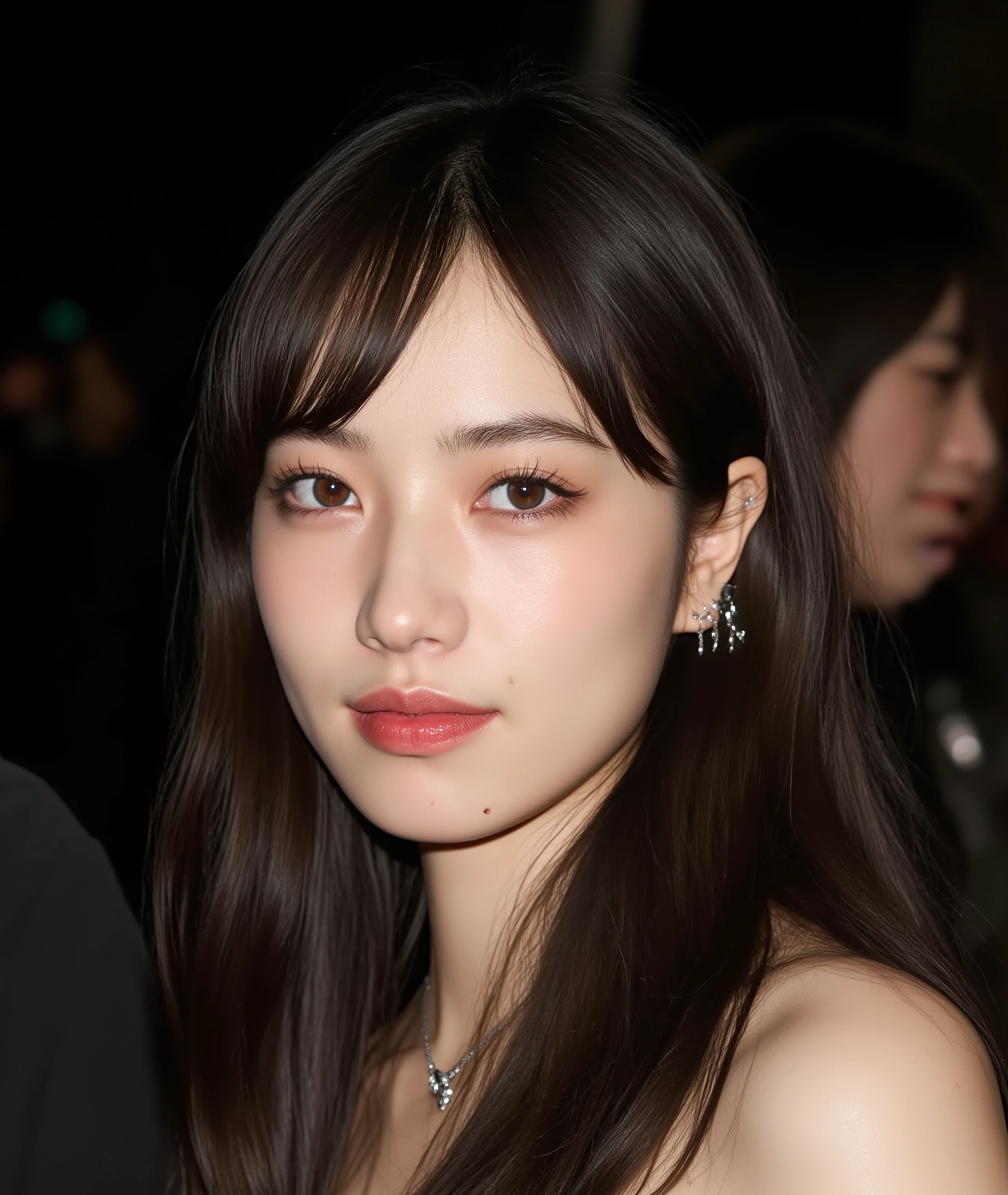 The image is a close-up photograph of a young Asian woman with a fair complexion. Her face dominates the frame, occupying the majority of the image, with her eyes, nose, and lips prominently visible. She has large, almond-shaped eyes with long, thick eyelashes, and her skin is smooth and soft. Her lips are full and slightly parted, revealing a natural, rosy hue. She wears a small silver stud earring in her left ear, adding a subtle touch of personal style.
Her hair is long, dark brown with subtle highlights, and it partially covers her forehead, giving a slightly tousled, casual look. The background is out of focus, but it appears to be a dark, possibly indoor setting, which emphasizes her face and provides a stark contrast to her light complexion. She is wearing a black garment, possibly a top or a sleeve, which is visible at the bottom of the image. The overall mood of the photograph is intimate and personal, capturing a candid moment with a natural, unfiltered aesthetic.