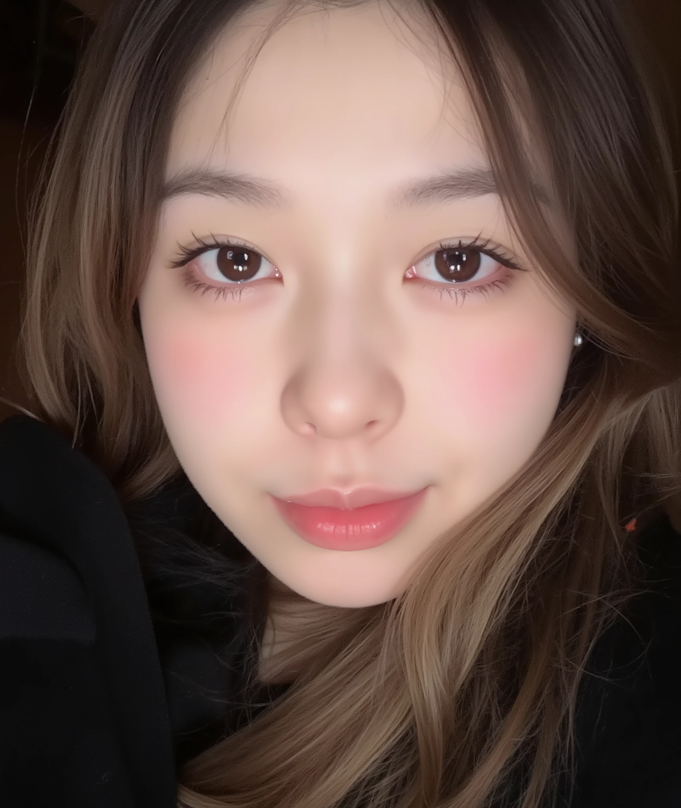 The image is a close-up photograph of a young Asian woman with a fair complexion. Her face dominates the frame, occupying the majority of the image, with her eyes, nose, and lips prominently visible. She has large, almond-shaped eyes with long, thick eyelashes, and her skin is smooth and soft. Her lips are full and slightly parted, revealing a natural, rosy hue. She wears a small silver stud earring in her left ear, adding a subtle touch of personal style.

Her hair is long, dark brown with subtle highlights, and it partially covers her forehead, giving a slightly tousled, casual look. The background is out of focus, but it appears to be a dark, possibly indoor setting, which emphasizes her face and provides a stark contrast to her light complexion. She is wearing a black garment, possibly a top or a sleeve, which is visible at the bottom of the image. The overall mood of the photograph is intimate and personal, capturing a candid moment with a natural, unfiltered aesthetic.