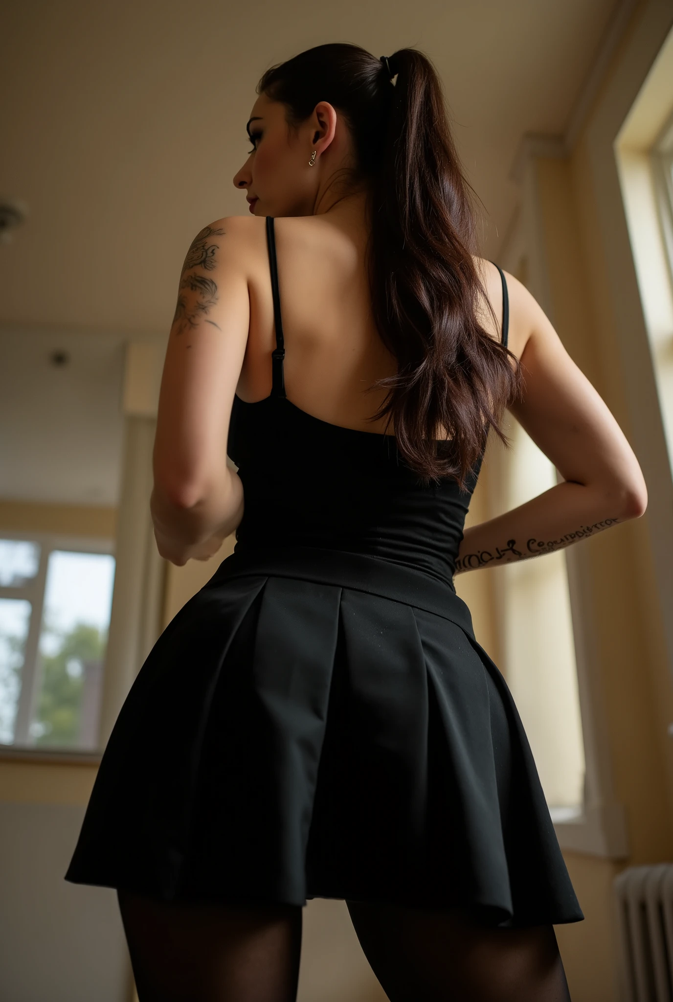 imposing rear-angle portrait of domineering European woman with stout powerful build, wide hips and thick strong legs in authoritative stance, minimal head turn revealing just a glimpse of stern profile with intense sidelong glare, wavy dark brown hair inspired by rockabilly style and tied into long full ponytail, fitted black pleated skirt with sheer black tights showcasing her substantial curves, black tank top with thin straps along her broad back, small tattoos on arms, back deliberately arched to assert dominance while emphasizing her full rounded buttocks, shot from extremely low submissive angle with ceiling visible beyond her towering form, bright daylight creating dramatic shadows that accentuate her commanding presence