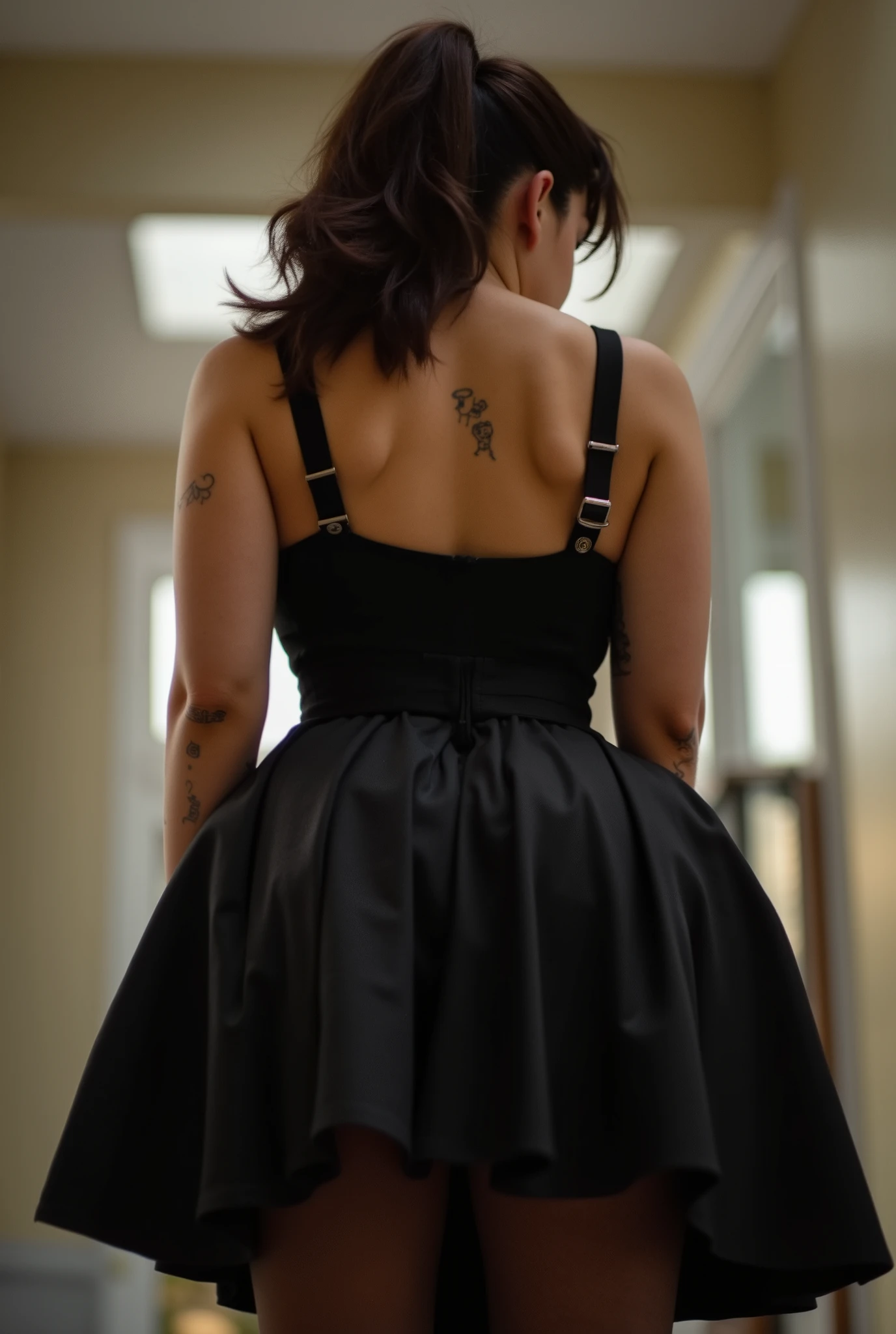 imposing rear-angle portrait of domineering European woman with stout powerful build, wide hips and thick strong legs in authoritative stance, minimal head turn revealing just a glimpse of stern profile with intense sidelong glare, wavy dark brown hair inspired by rockabilly style and tied into long full ponytail, fitted black pleated skirt with sheer black tights showcasing her substantial curves, black tank top with thin straps along her broad back, small tattoos on arms, back deliberately arched to assert dominance while emphasizing her full rounded buttocks, shot from extremely low submissive angle with ceiling visible beyond her towering form, bright daylight creating dramatic shadows that accentuate her commanding presence