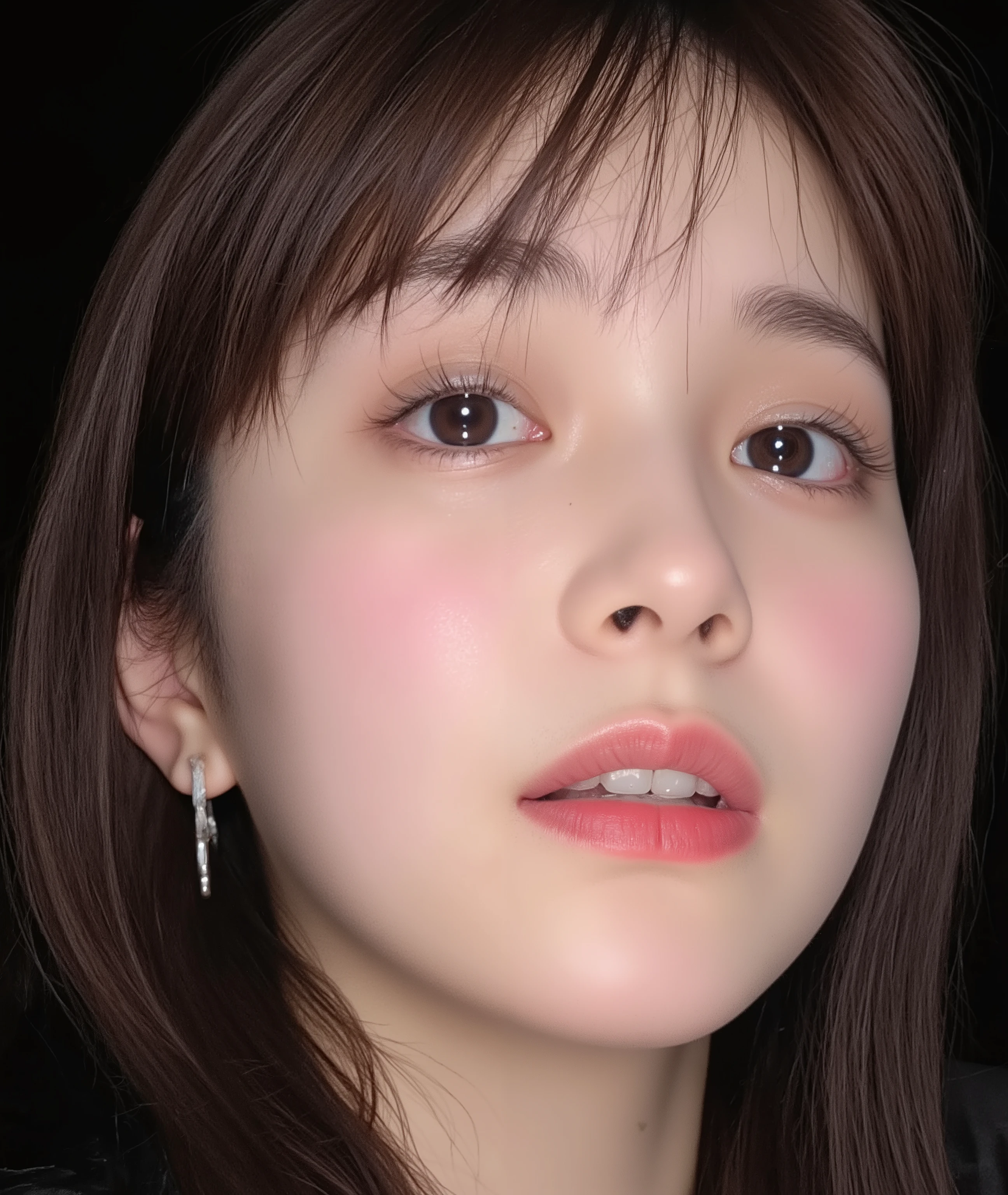 The image is a close-up photograph of a young Asian woman with a fair complexion. Her face dominates the frame, occupying the majority of the image, with her eyes, nose, and lips prominently visible. She has large, almond-shaped eyes with long, thick eyelashes, and her skin is smooth and soft. Her lips are full and slightly parted, revealing a natural, rosy hue. She wears a small silver stud earring in her left ear, adding a subtle touch of personal style.
Her hair is long, dark brown with subtle highlights, and it partially covers her forehead, giving a slightly tousled, casual look. The background is out of focus, but it appears to be a dark, possibly indoor setting, which emphasizes her face and provides a stark contrast to her light complexion. She is wearing a black garment, possibly a top or a sleeve, which is visible at the bottom of the image. The overall mood of the photograph is intimate and personal, capturing a candid moment with a natural, unfiltered aesthetic.