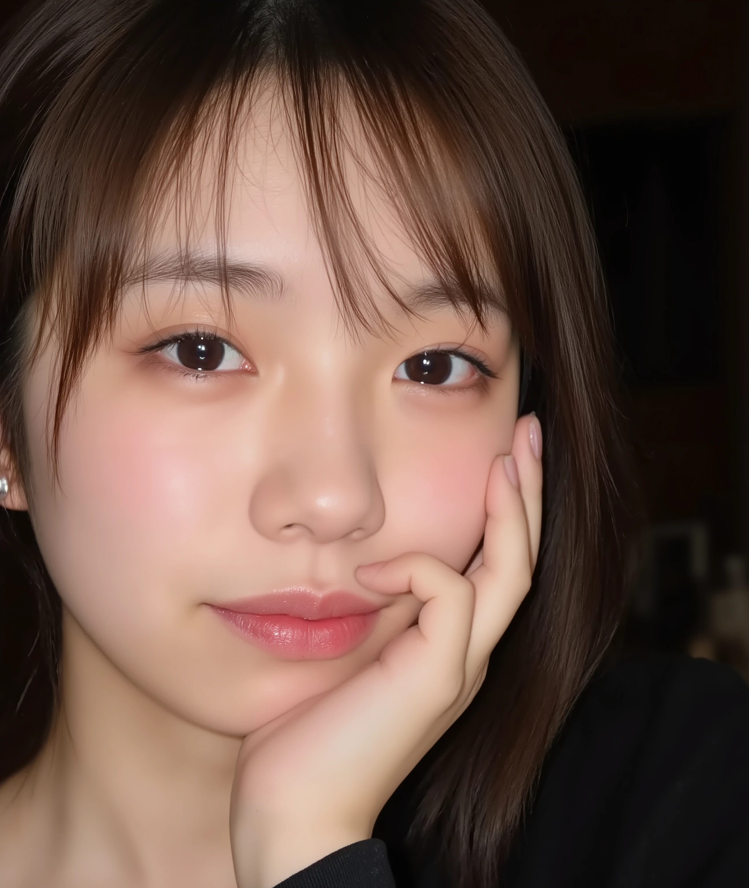 The image is a close-up photograph of a young Asian woman with a fair complexion. Her face dominates the frame, occupying the majority of the image, with her eyes, nose, and lips prominently visible. She has large, almond-shaped eyes with long, thick eyelashes, and her skin is smooth and soft. Her lips are full and slightly parted, revealing a natural, rosy hue. She wears a small silver stud earring in her left ear, adding a subtle touch of personal style.

Her hair is long, dark brown with subtle highlights, and it partially covers her forehead, giving a slightly tousled, casual look. The background is out of focus, but it appears to be a dark, possibly indoor setting, which emphasizes her face and provides a stark contrast to her light complexion. She is wearing a black garment, possibly a top or a sleeve, which is visible at the bottom of the image. The overall mood of the photograph is intimate and personal, capturing a candid moment with a natural, unfiltered aesthetic.