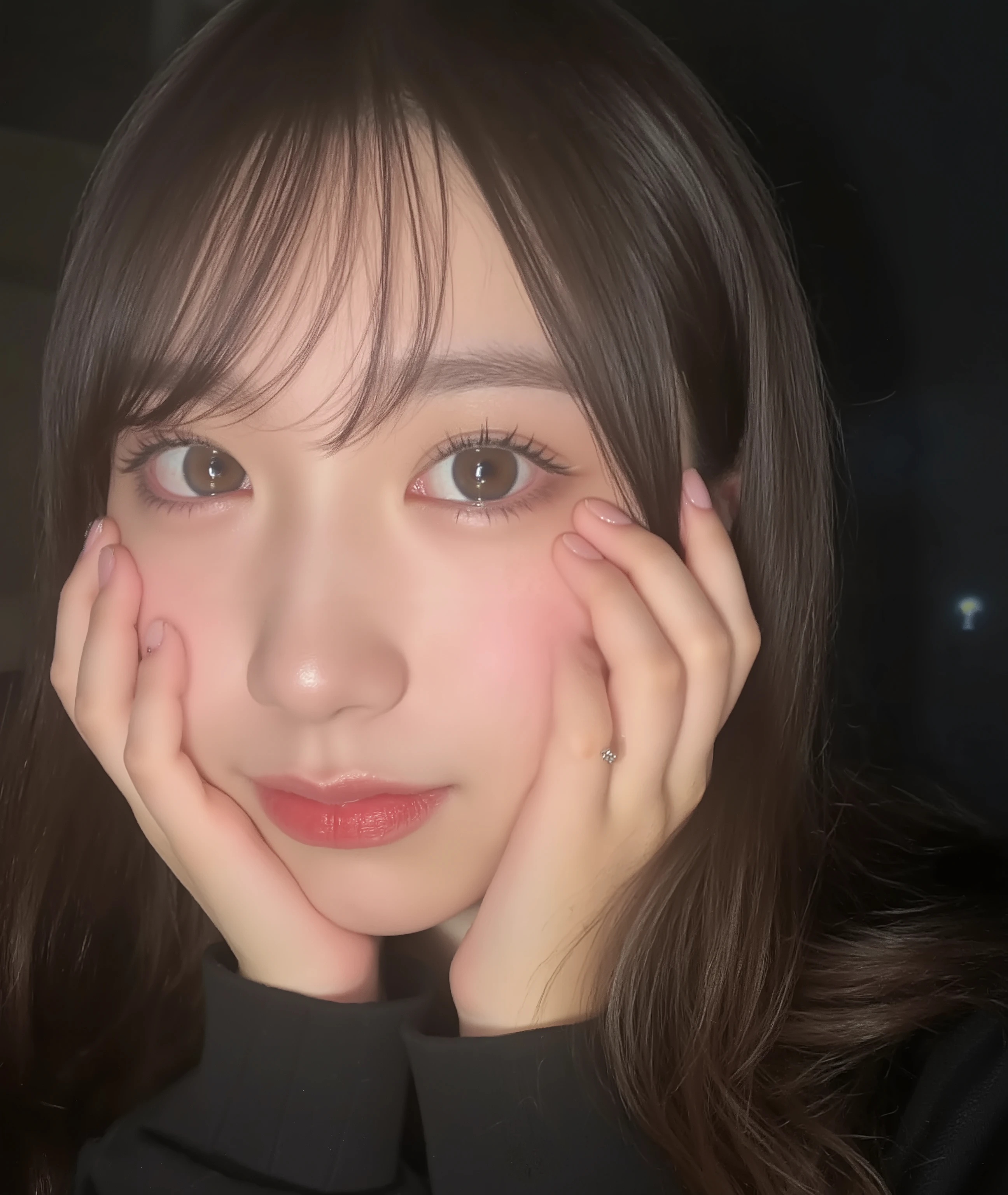 The image is a close-up photograph of a young Asian woman with a fair complexion. Her face dominates the frame, occupying the majority of the image, with her eyes, nose, and lips prominently visible. She has large, almond-shaped eyes with long, thick eyelashes, and her skin is smooth and soft. Her lips are full and slightly parted, revealing a natural, rosy hue. She wears a small silver stud earring in her left ear, adding a subtle touch of personal style.

Her hair is long, dark brown with subtle highlights, and it partially covers her forehead, giving a slightly tousled, casual look. The background is out of focus, but it appears to be a dark, possibly indoor setting, which emphasizes her face and provides a stark contrast to her light complexion. She is wearing a black garment, possibly a top or a sleeve, which is visible at the bottom of the image. The overall mood of the photograph is intimate and personal, capturing a candid moment with a natural, unfiltered aesthetic.