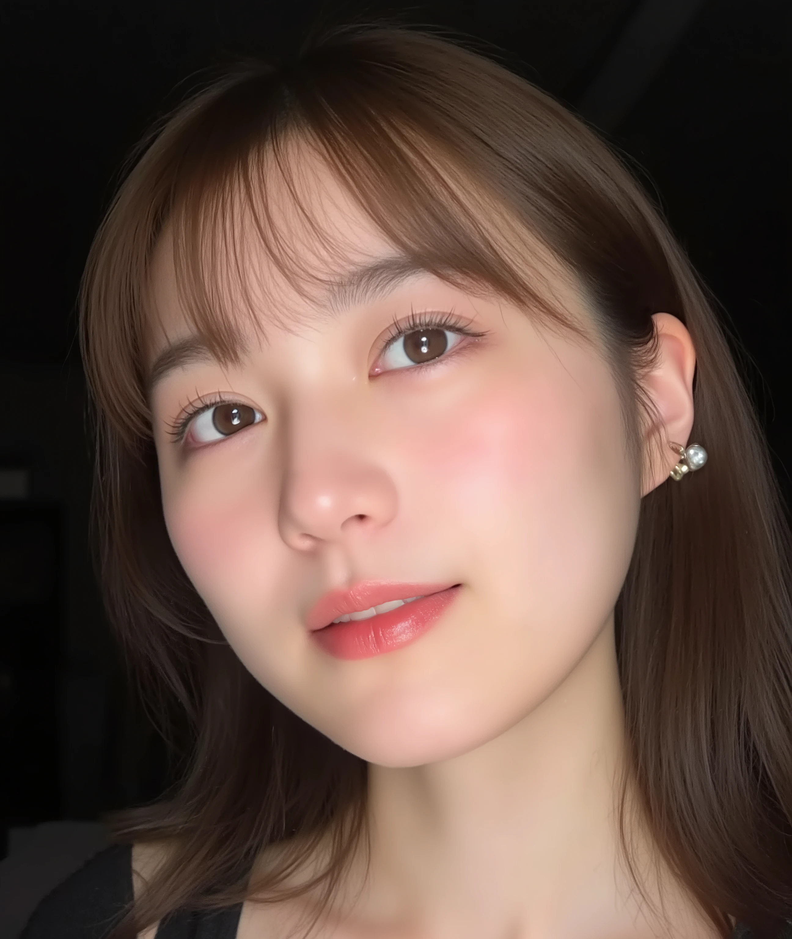 The image is a close-up photograph of a young Asian woman with a fair complexion. Her face dominates the frame, occupying the majority of the image, with her eyes, nose, and lips prominently visible. She has large, almond-shaped eyes with long, thick eyelashes, and her skin is smooth and soft. Her lips are full and slightly parted, revealing a natural, rosy hue. She wears a small silver stud earring in her left ear, adding a subtle touch of personal style.
Her hair is long, dark brown with subtle highlights, and it partially covers her forehead, giving a slightly tousled, casual look. The background is out of focus, but it appears to be a dark, possibly indoor setting, which emphasizes her face and provides a stark contrast to her light complexion. She is wearing a black garment, possibly a top or a sleeve, which is visible at the bottom of the image. The overall mood of the photograph is intimate and personal, capturing a candid moment with a natural, unfiltered aesthetic.