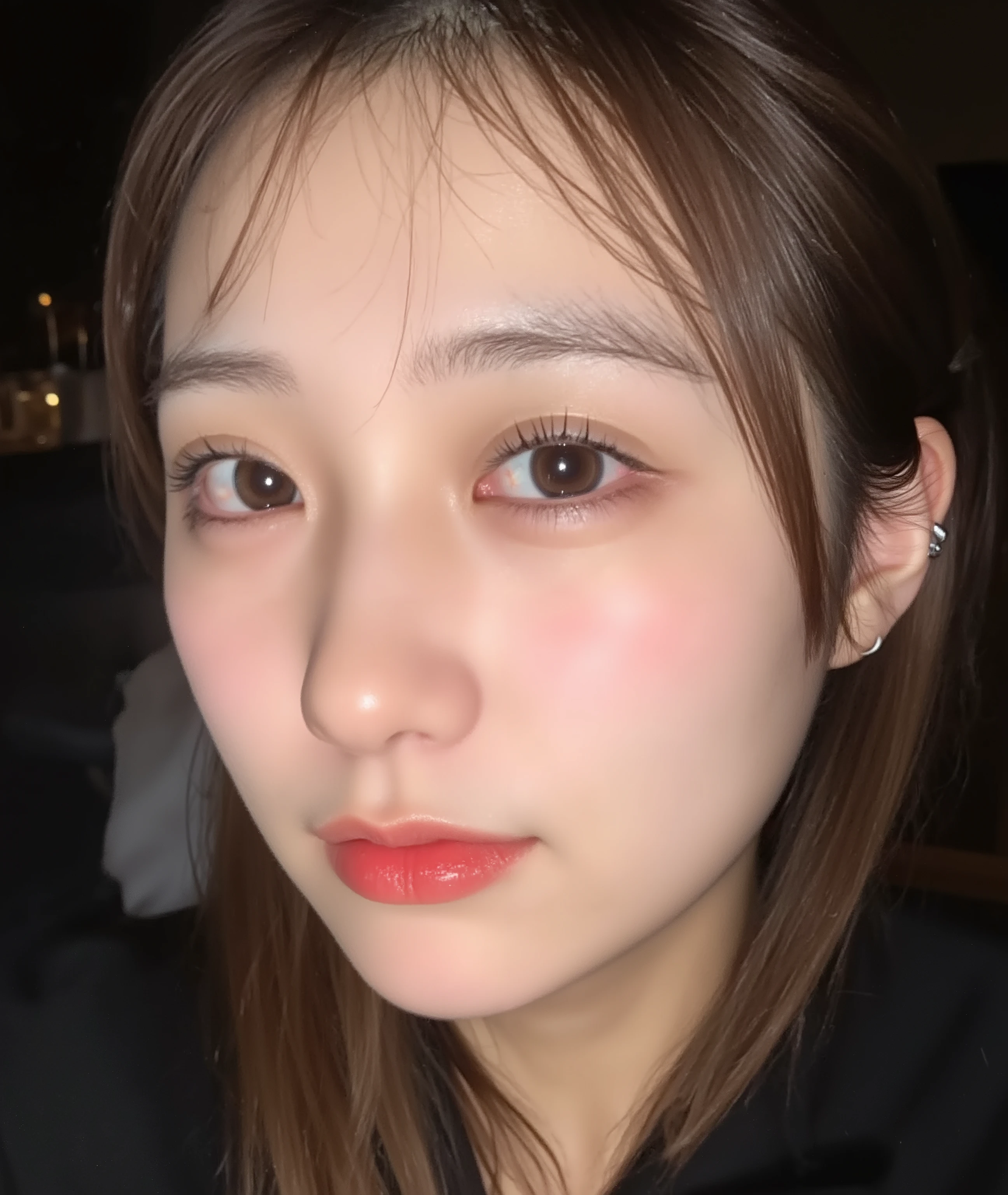 The image is a close-up photograph of a young Asian woman with a fair complexion. Her face dominates the frame, occupying the majority of the image, with her eyes, nose, and lips prominently visible. She has large, almond-shaped eyes with long, thick eyelashes, and her skin is smooth and soft. Her lips are full and slightly parted, revealing a natural, rosy hue. She wears a small silver stud earring in her left ear, adding a subtle touch of personal style.
Her hair is long, dark brown with subtle highlights, and it partially covers her forehead, giving a slightly tousled, casual look. The background is out of focus, but it appears to be a dark, possibly indoor setting, which emphasizes her face and provides a stark contrast to her light complexion. She is wearing a black garment, possibly a top or a sleeve, which is visible at the bottom of the image. The overall mood of the photograph is intimate and personal, capturing a candid moment with a natural, unfiltered aesthetic.
