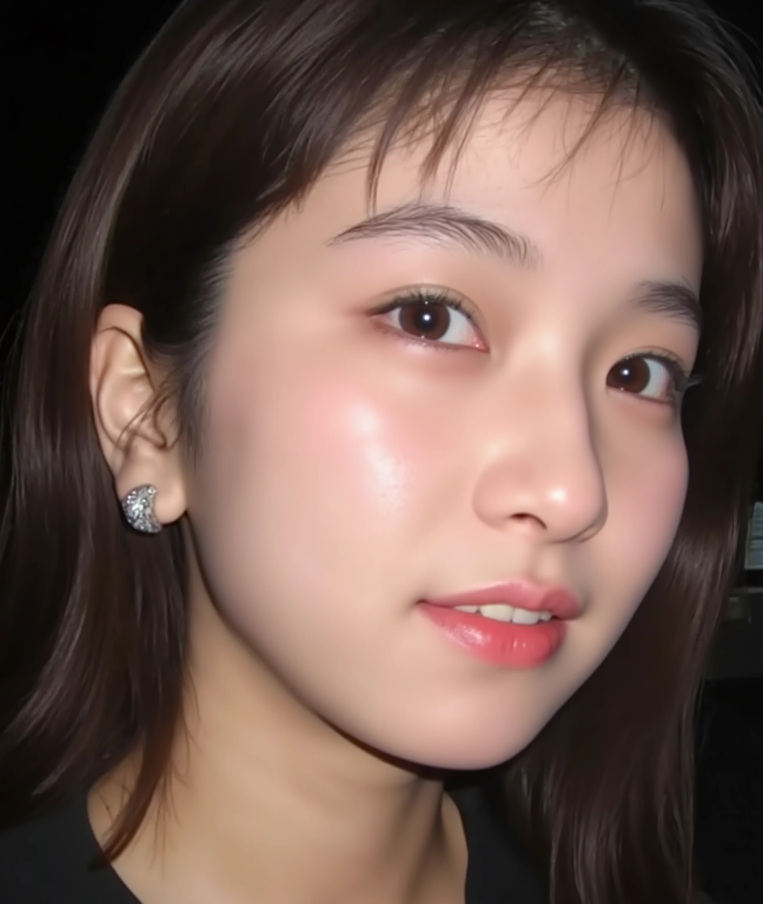 The image is a close-up photograph of a young Asian woman with a fair complexion. Her face dominates the frame, occupying the majority of the image, with her eyes, nose, and lips prominently visible. She has large, almond-shaped eyes with long, thick eyelashes, and her skin is smooth and soft. Her lips are full and slightly parted, revealing a natural, rosy hue. She wears a small silver stud earring in her left ear, adding a subtle touch of personal style.
Her hair is long, dark brown with subtle highlights, and it partially covers her forehead, giving a slightly tousled, casual look. The background is out of focus, but it appears to be a dark, possibly indoor setting, which emphasizes her face and provides a stark contrast to her light complexion. She is wearing a black garment, possibly a top or a sleeve, which is visible at the bottom of the image. The overall mood of the photograph is intimate and personal, capturing a candid moment with a natural, unfiltered aesthetic.