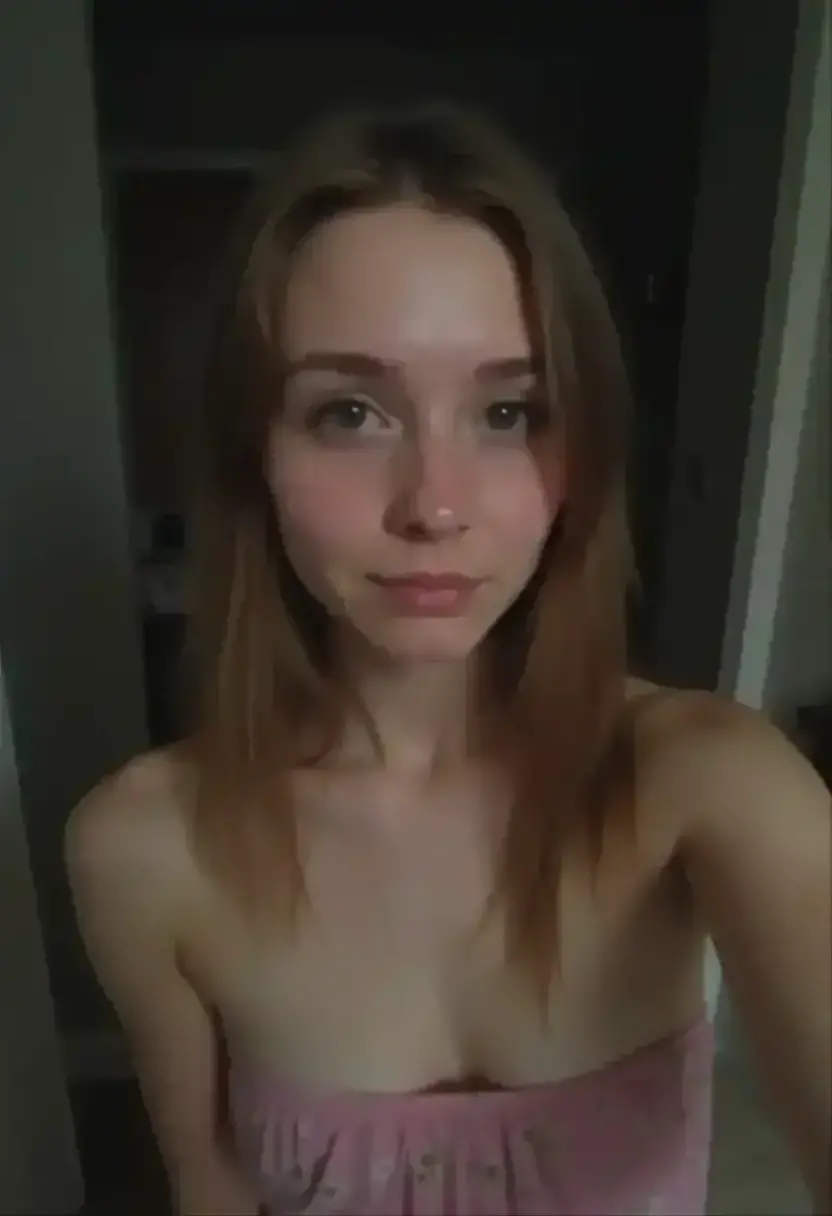 The image is a high-resolution photograph of a nude young woman, likely in her early twenties, with a slender, petite physique and light skin. She has straight, medium-length light brown hair with bangs that frame her face. She is smiling softly, giving a casual, friendly expression. Her breasts are small to medium-sized, and she has a small, natural-looking pubic area with visible pubic hair.