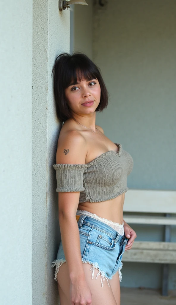 A young, light-skinned woman, likely in her teens or early twenties, is positioned slightly off-center, leaning against a light gray concrete pillar.  She is facing the camera, and her expression is neutral.  Her hair is short and dark brown.  She has a small heart-shaped tattoo on her shoulder.  She is wearing a random color, off-the-shoulder, crop top with a slightly gathered, textured bodice and short, puff sleeves.  She is also wearing light blue denim jeans and sheer, off-white lace panties.  Her body build is curvy. The environmental context is an outdoor setting, with a light gray, textured wall forming the backdrop. A light-colored park bench is visible in the background.  The lighting is soft and natural, creating a bright and somewhat airy atmosphere. The composition is straightforward and slightly angled, with the subject's body positioned in a casual, yet slightly suggestive pose. The overall aesthetic is stylish and trendy,  in a contemporary casual style. The colors are muted A small light fixture is seen on the upper left of the image.
