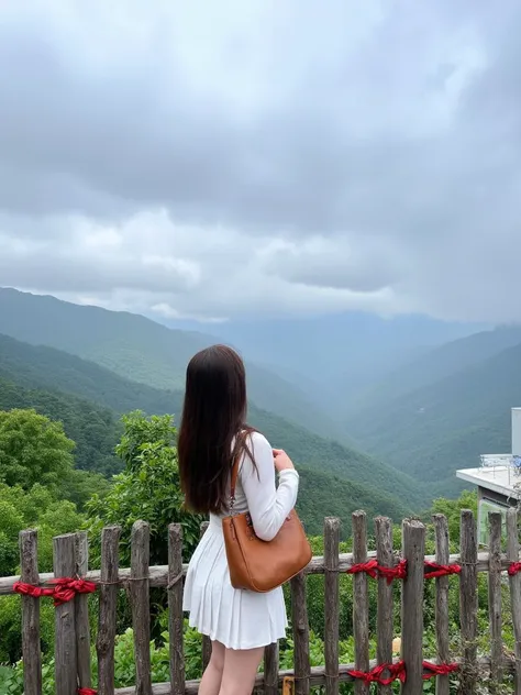 The image is a photograph capturing a serene mountainous landscape. The primary subject is a young woman standing with her back to the camera, gazing at the vast, mist-shrouded mountains below. She has long, straight brown hair cascading down her back and is dressed in a white, long-sleeve top and matching short, pleated skirt. A large, brown leather handbag hangs over her right shoulder. Her skin is fair, and she appears to be of East Asian descent. 

The background showcases a panoramic view of dense, green mountain ranges stretching into the distance, with low-lying clouds adding a mystical touch. The sky above is filled with dramatic, overcast clouds, suggesting a changeable weather pattern. In the foreground, a rustic wooden fence runs horizontally, adorned with red ribbons tied to it, possibly indicating a memorial or cultural significance. To the right, a modern building with a flat roof and white exterior sits partially obscured by vegetation, hinting at a blend of traditional and contemporary elements in the setting. The overall scene exudes tranquility and natural beauty.