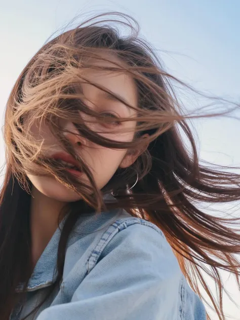 This photograph captures a close-up, low-angle view of a young woman with long, dark brown hair that is blowing gently in the wind. She is of East Asian descent, with smooth, fair skin and a natural, youthful appearance. Her face is partially obscured by her hair, which frames her face and partially covers her eyes. The texture of her hair appears silky and slightly tousled, giving it a natural, windblown look. She is wearing a light blue denim jacket, which is visible at the top of the image, with its fabric appearing soft and slightly worn. The background is a clear, pale blue sky, suggesting that the photo was taken outdoors during the day, possibly in the early morning or late afternoon, given the soft, golden light that enhances her features. The image exudes a sense of freedom and natural beauty, with the focus on the woman's face and hair, rather than her body or surroundings. The overall mood is serene and contemplative, emphasizing the beauty of the moment rather than any specific action or event.