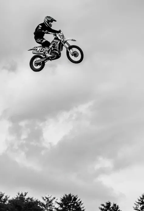 Motocross Mid-Air Majesty, black and white analog photograph, motocross rider, dirt bike number 39, mid-air jump, motocross helmet, goggles, leather jacket, action shot, dynamic pose, clear sky, outdoor sport, motorcycle sport, photography techniques motion freeze, observer in background, dirt terrain, blurred background, sports event, vintage motorcycle gear, speed impression, sense of danger, high contrast image, text "HONDA".