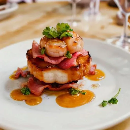 A plate of perfectly seared scallops wrapped in crispy bacon, garnished with fresh parsley and served with a side of garlic butter sauce. The scallops are tender and slightly caramelized on the edges, with the bacon adding a savory crunch. The dish is served on a white plate with a glass of chilled white wine.
