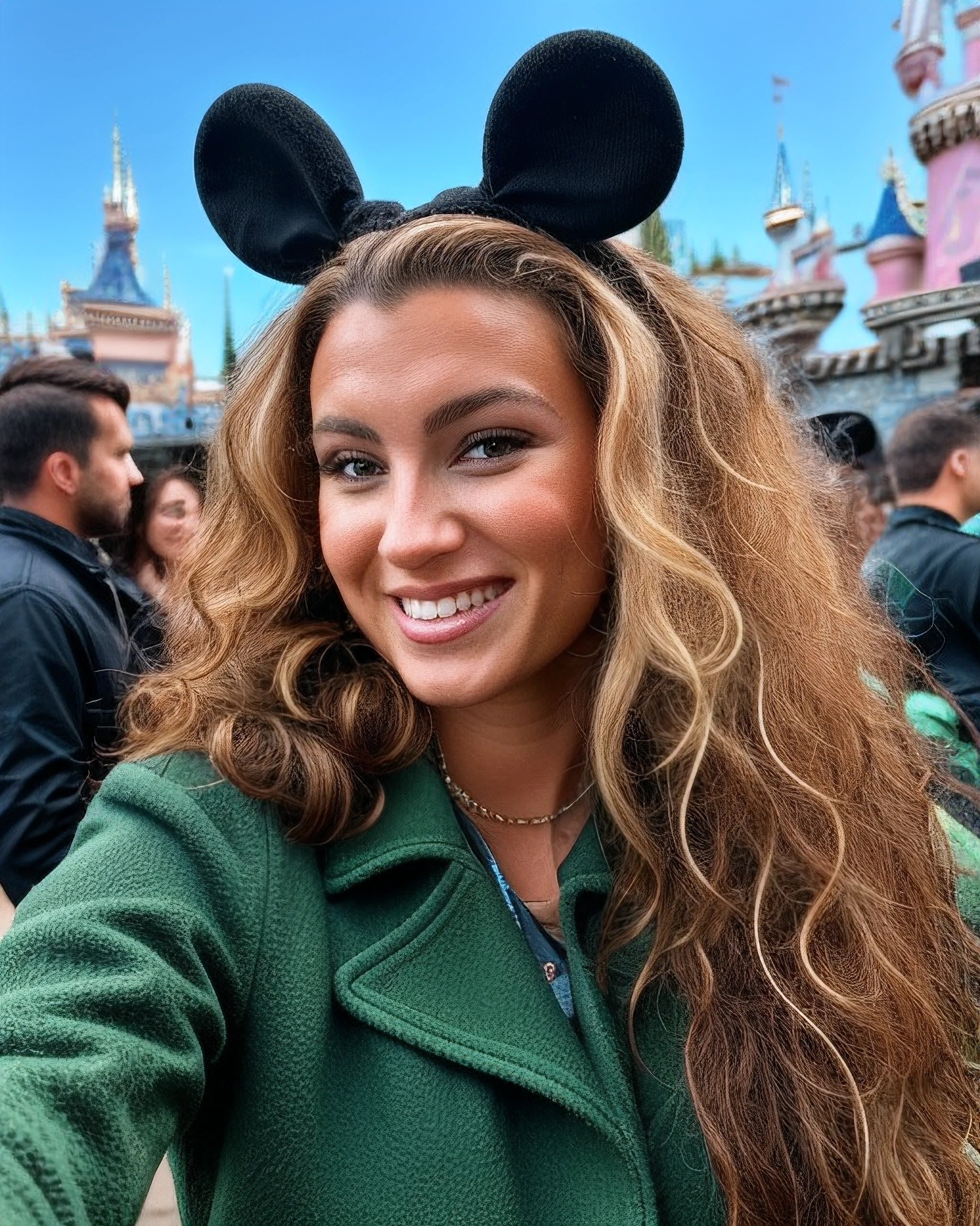 tori-k, close-up, (realistic), (hyperrealism), long hair, blonde highlights, happy, playful, (photorealistic:1.4), best quality, masterpiece, messy hair, curly hair,at disneyland, wearing mouse ears, green overcoat, handbag, necklace