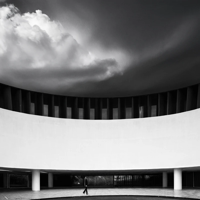 black & white, a solitary figure standing under a vast, circular open structure with a patterned roof revealing the cloudy sky, minimalist architecture, modern art style, high-contrast elements, dramatic lighting

